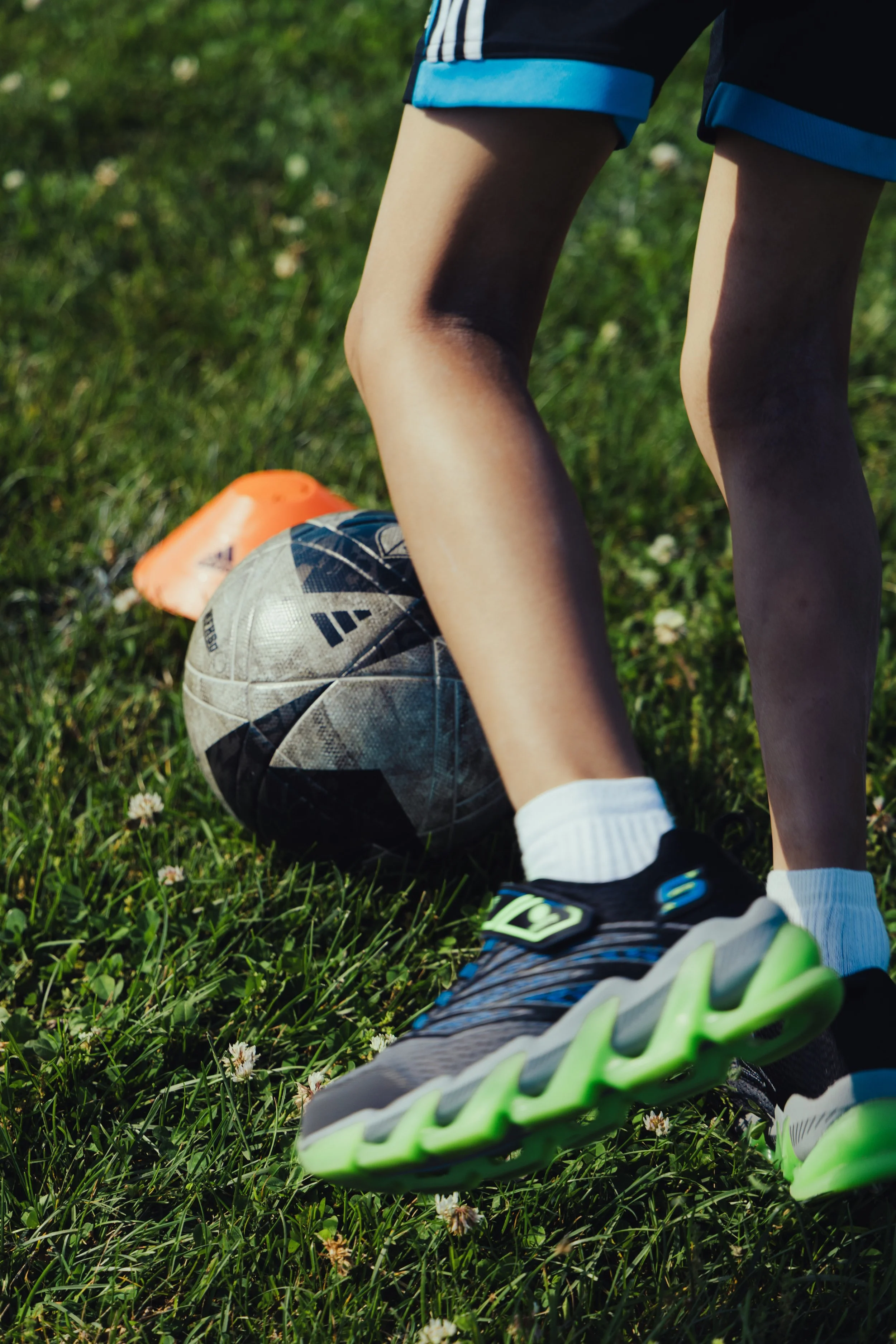 Close-up of a child in soccer gear lying on grass with one foot on a soccer ball and the other foot in a green and black athletic shoe; the child's knee and shorts are visible, and orange cones are in the background.