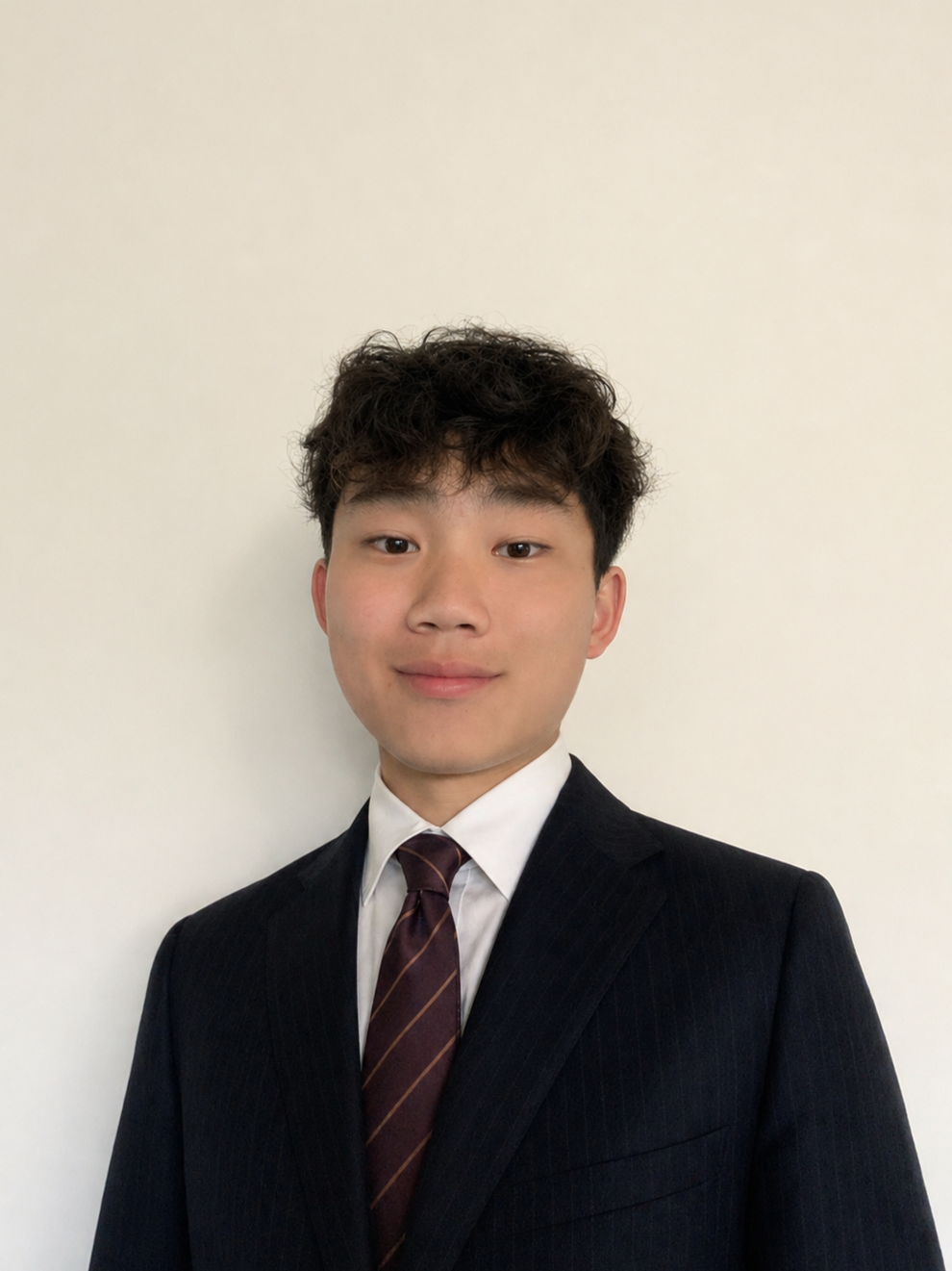 A young man in a black pinstripe suit, white shirt, and maroon striped tie standing against a plain off-white wall.