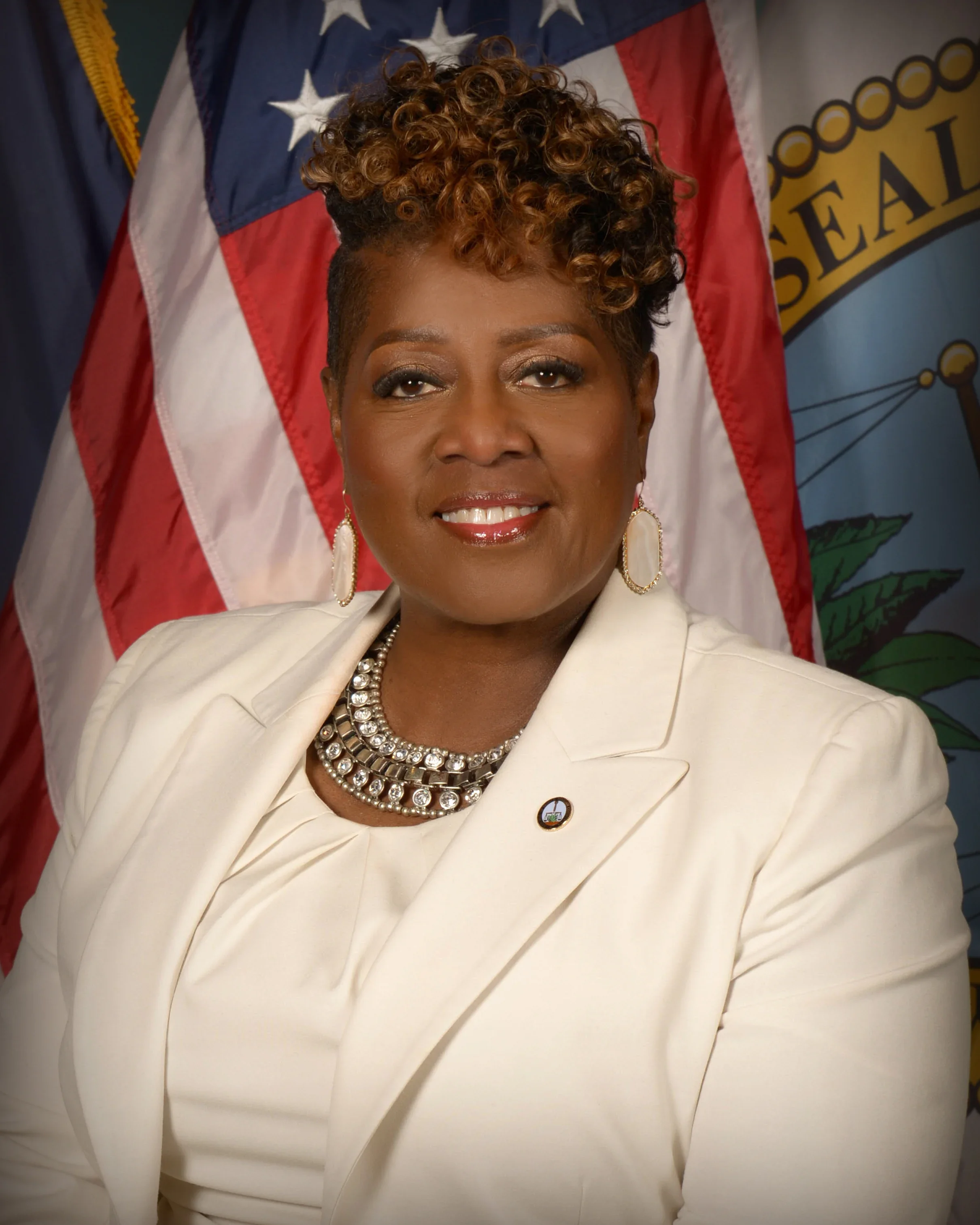 Professional woman in white suit with jewelry, standing in front of American flag and state seal.