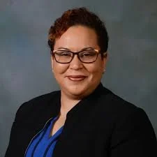 Portrait of a woman with short curly hair, glasses, wearing a black blazer and blue blouse, smiling against a gray background.