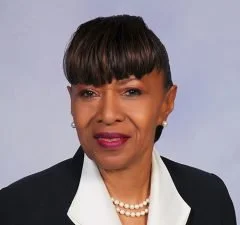 A woman with short black hair, wearing a white shirt, black blazer, pearl necklace, and pearl earrings against a light background.