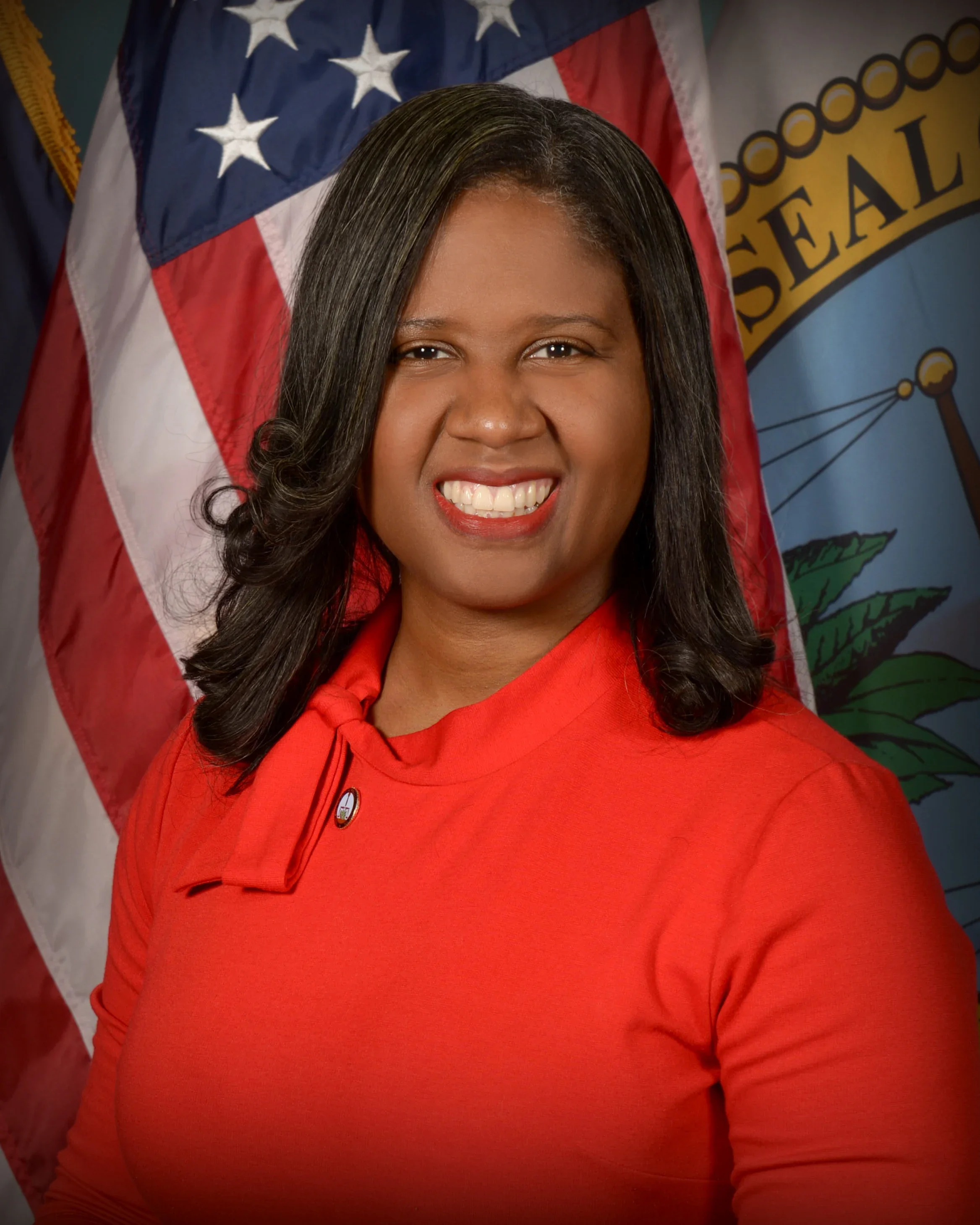 A woman with shoulder-length black hair smiling, wearing a red dress with a small pin, standing in front of American flags and a seal.