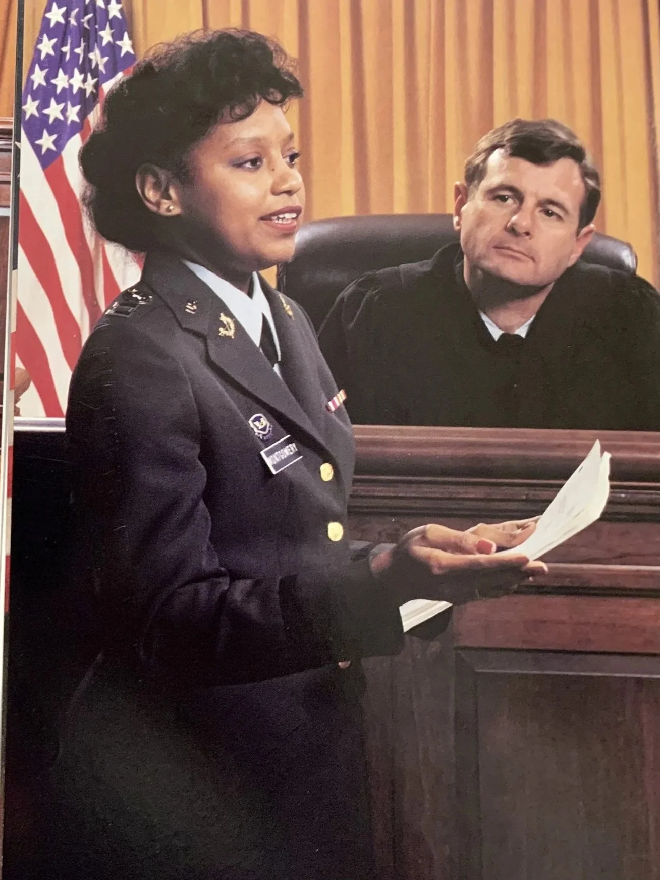 A woman in military uniform speaking in a courtroom, with a judge listening and looking on.
