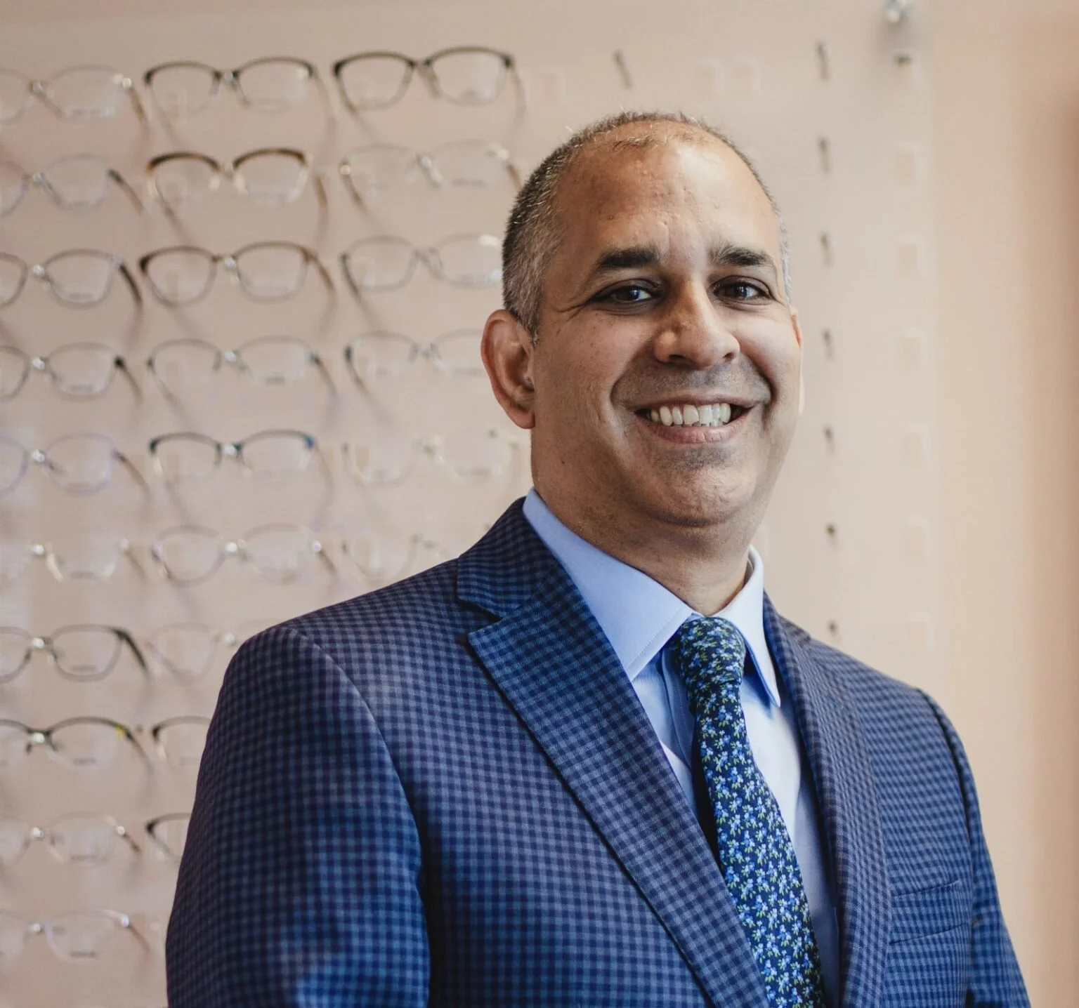 A smiling man in a blue checkered suit and patterned tie standing in front of a wall displaying multiple eyeglasses.