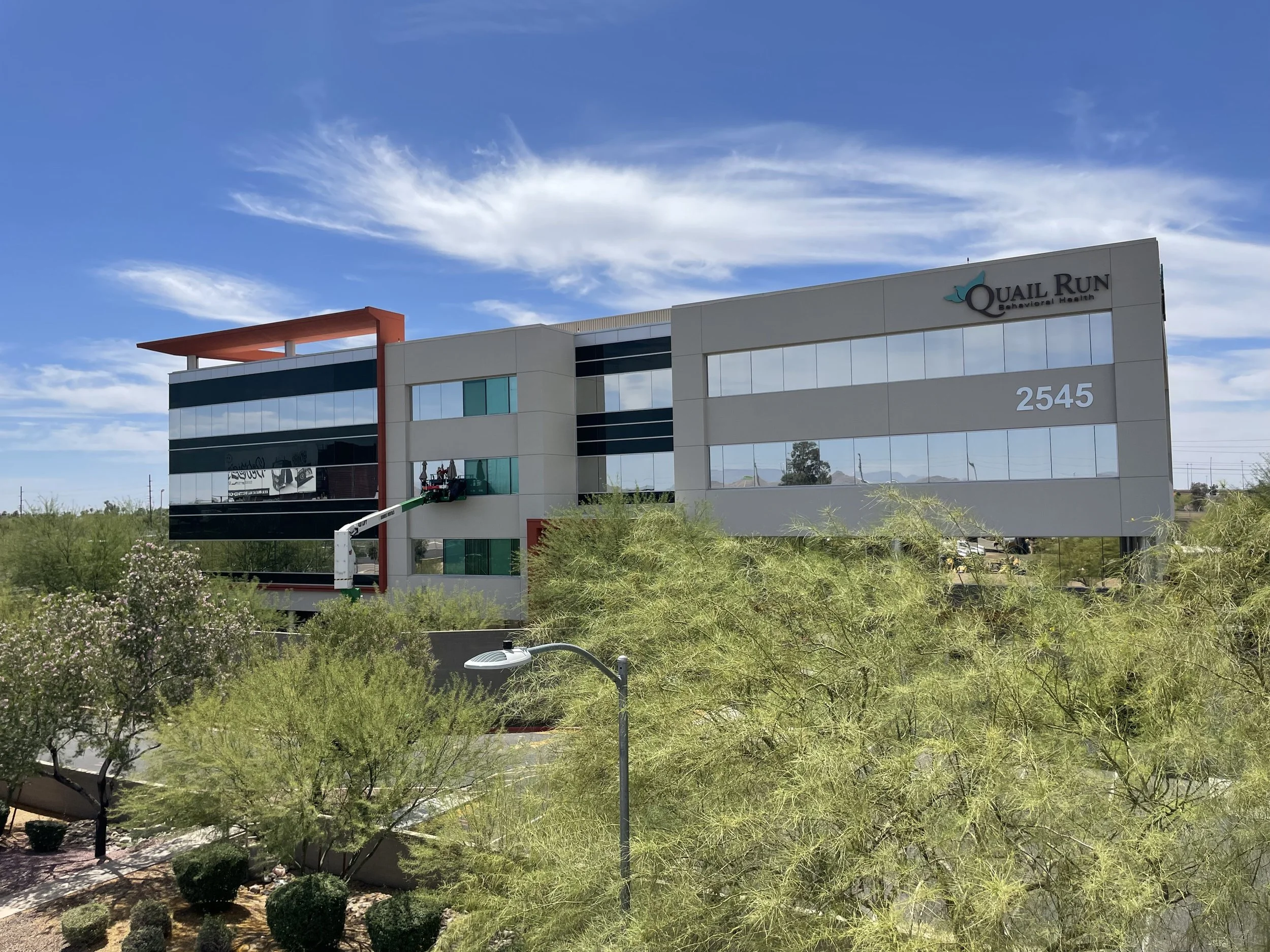 Exterior of a modern office building with the sign 'Quail Run Behavioral Health' and the street number 2545, surrounded by desert vegetation and trees, under a blue sky with some clouds.