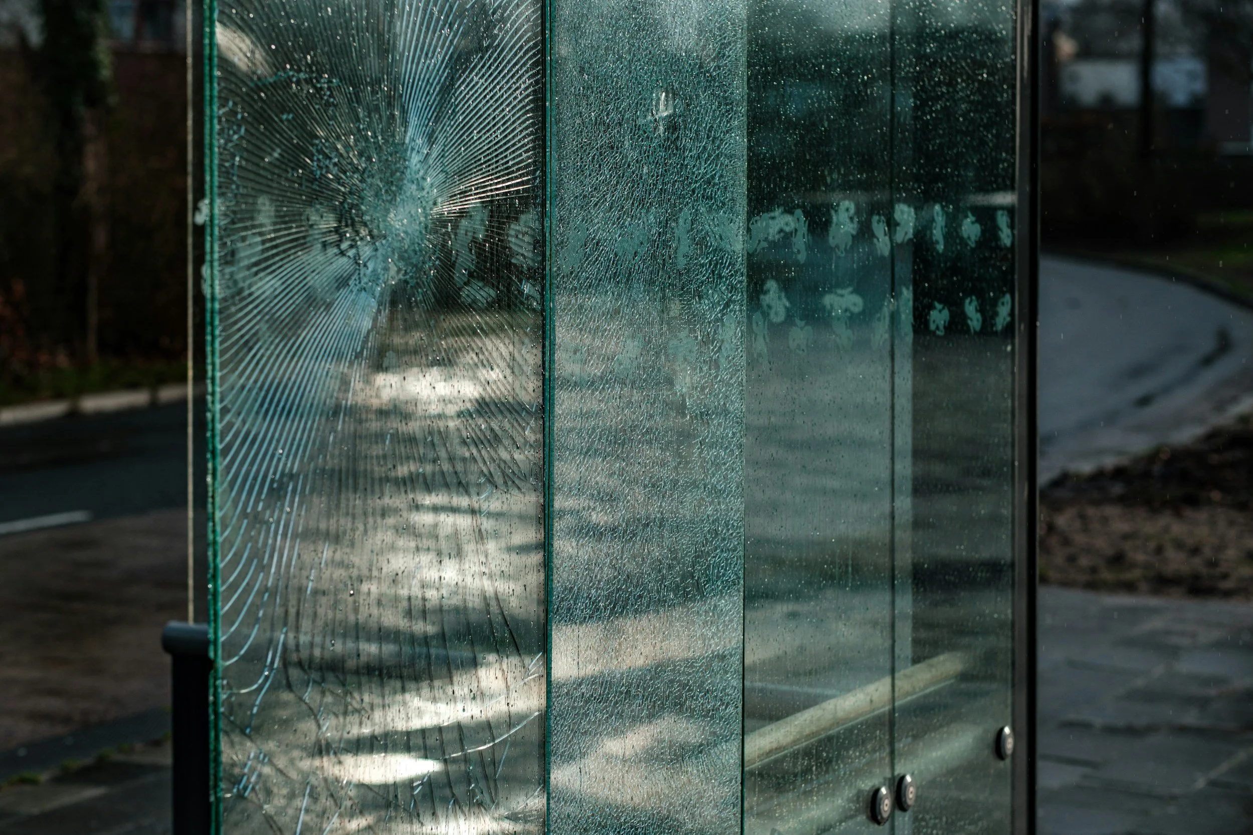 A glass panel with a large impact point and radiating cracks, likely from a thrown or struck object, with the background showing a street and trees.
