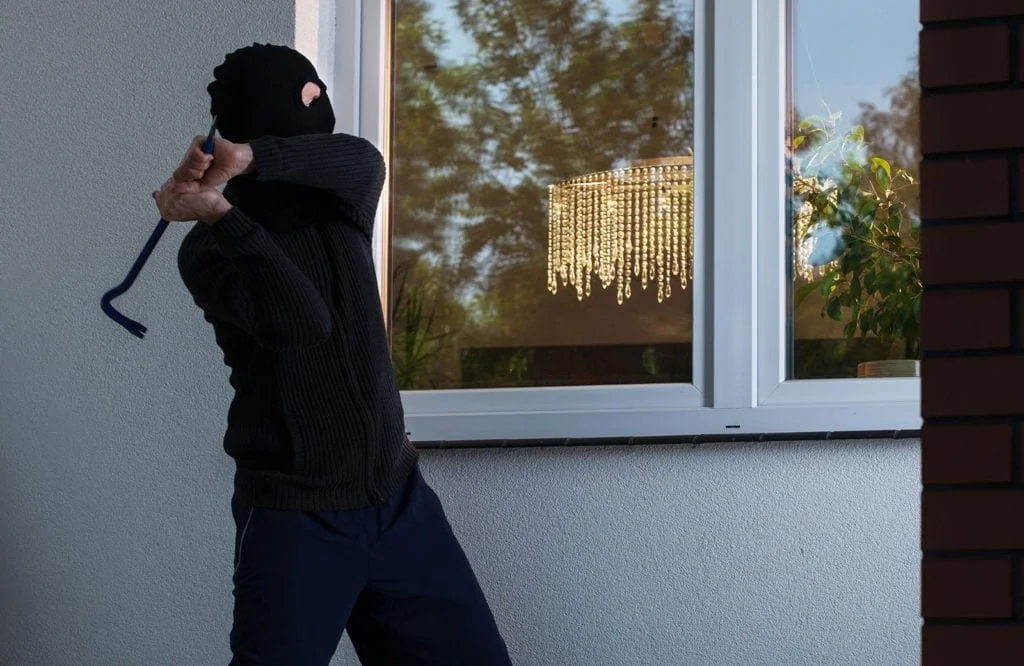 A person dressed as a burglar wearing a black ski mask and dark clothing is holding a crowbar, standing outside near a window.