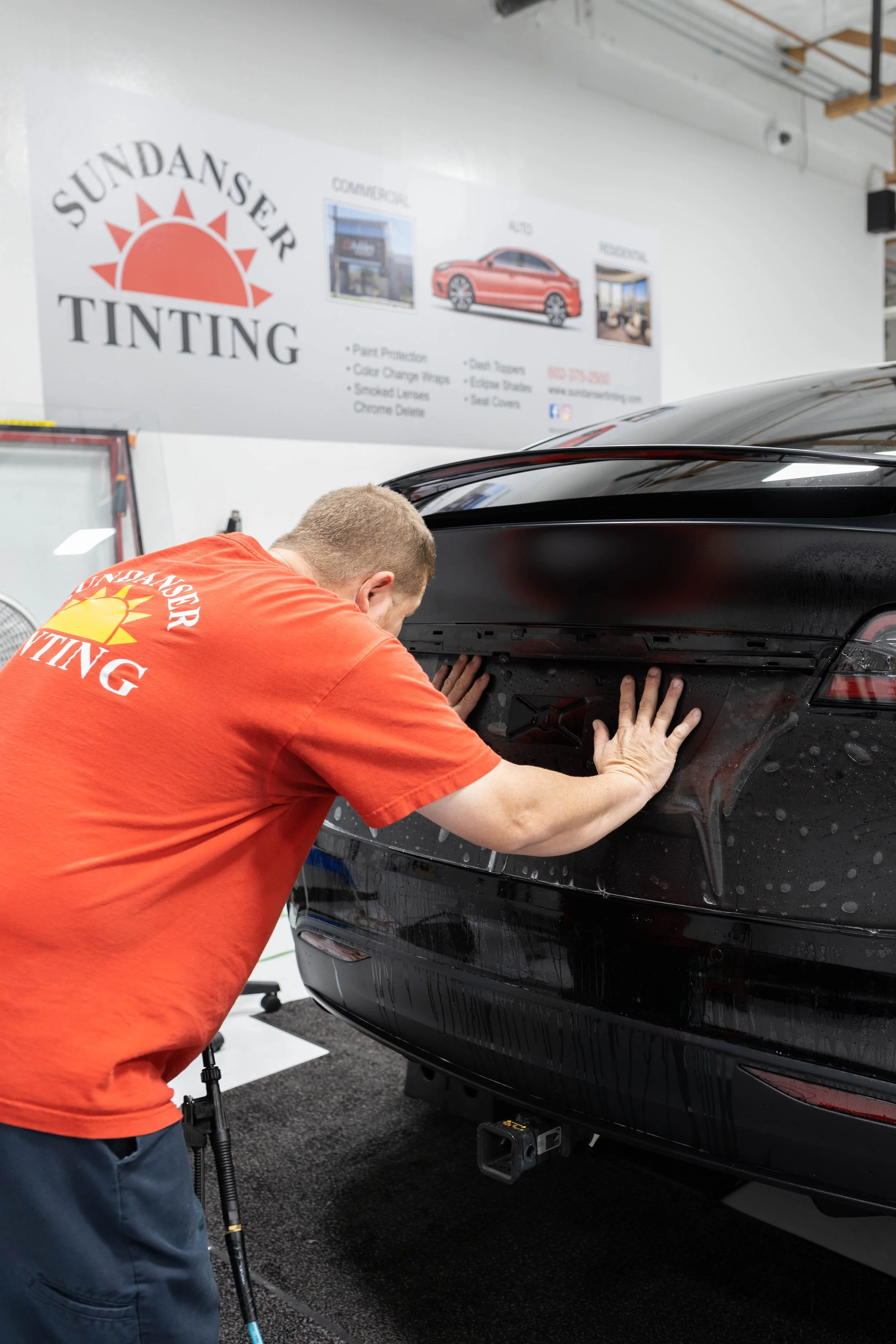 A person in an orange shirt with a 'Sundancer Tinting' logo on the back is working on a black car, possibly tinting the windows, inside a workshop with a 'Sundancer Tinting' sign on the wall.