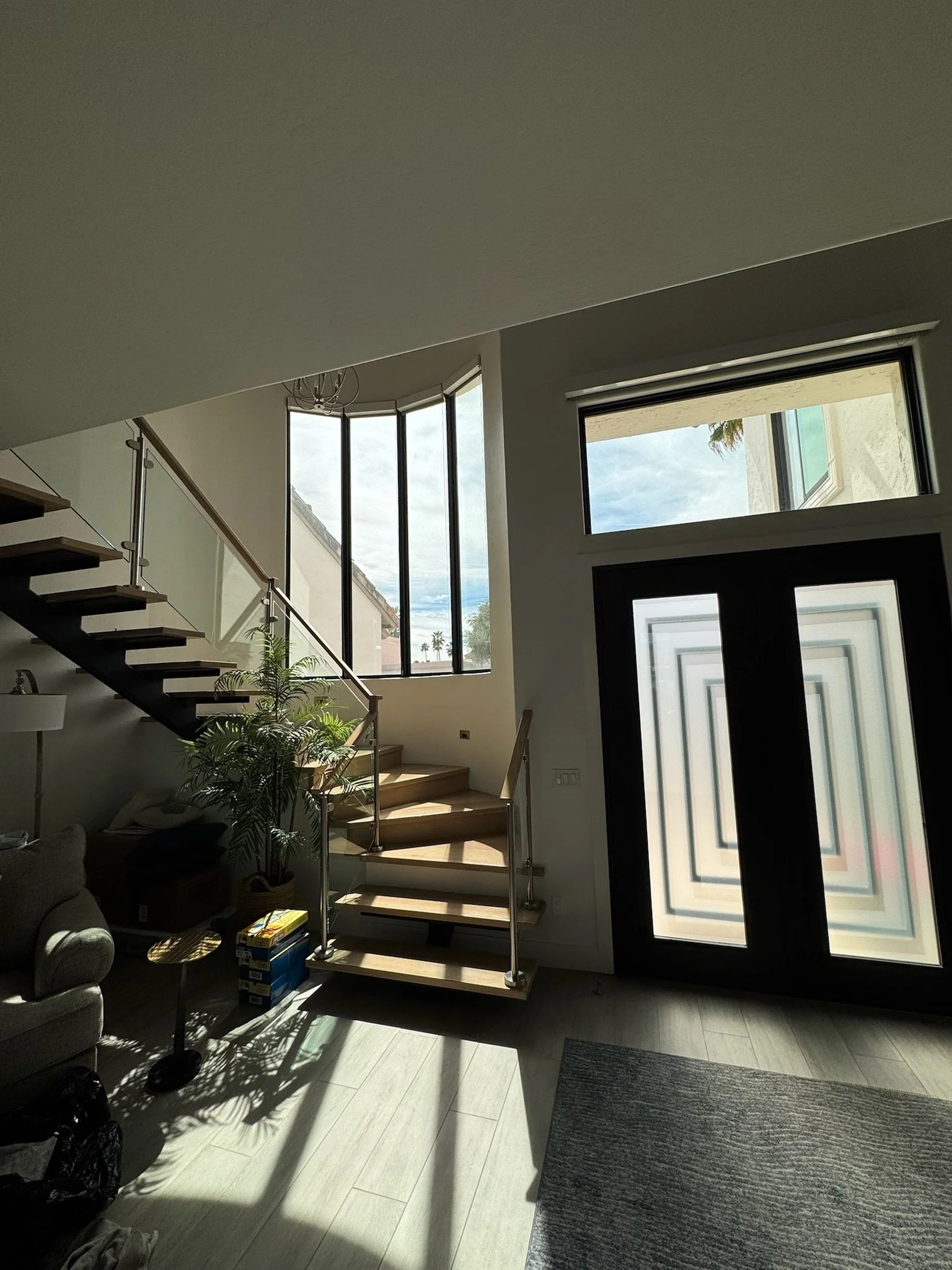 Interior of a modern house with a staircase, large windows, a door with stained glass, a standing lamp, a sofa, a small table, a plant, and a rug on light-colored flooring.