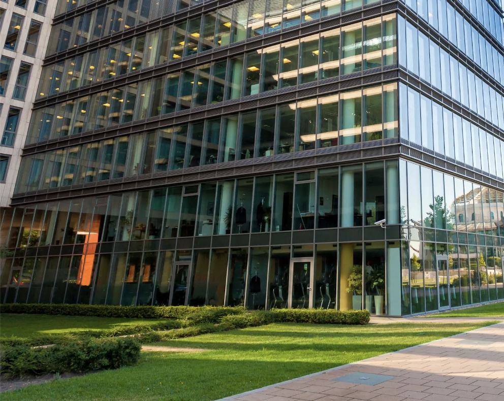 Exterior of a modern glass office building with reflections, grass, and sidewalk in the foreground.