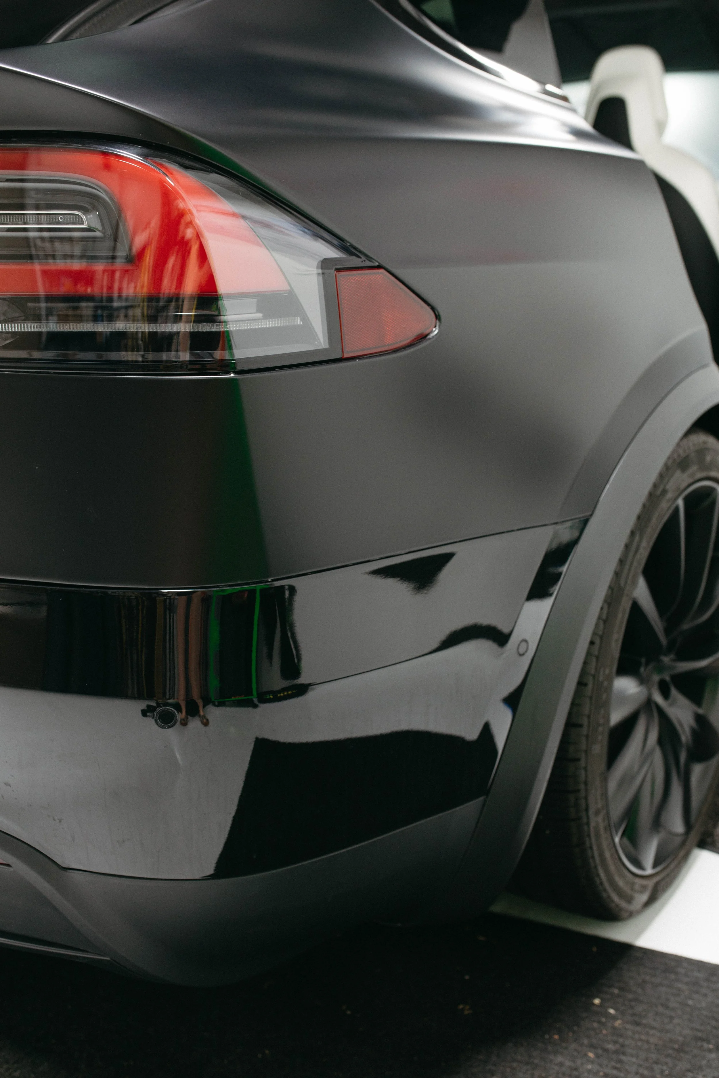 Close-up of the rear of a black electric car, showing the taillight and rear wheel, parked indoors.