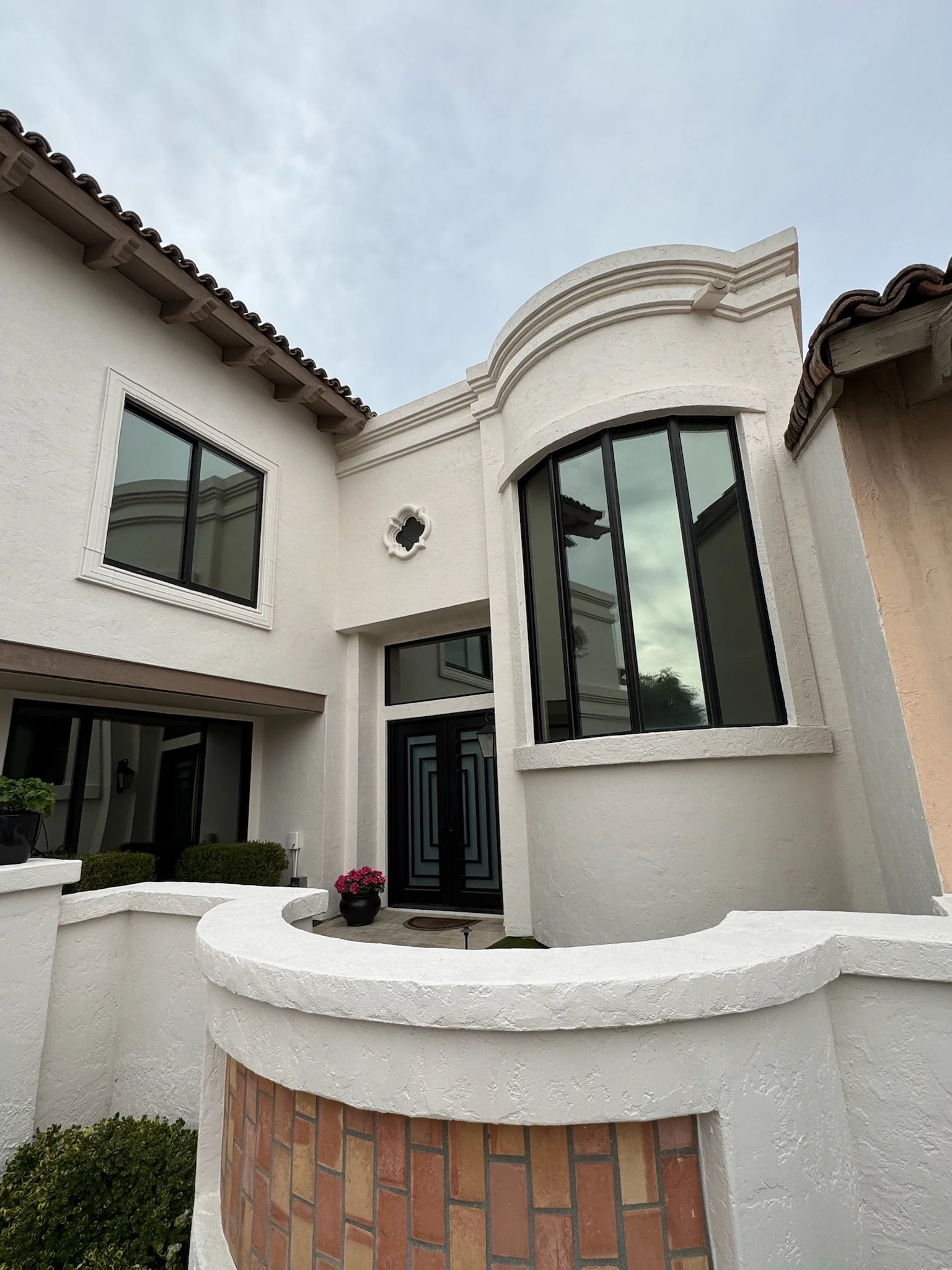 Front view of a modern house with white stucco exterior, large black-framed windows, decorative molding, and a small brick courtyard with potted plants and bushes.
