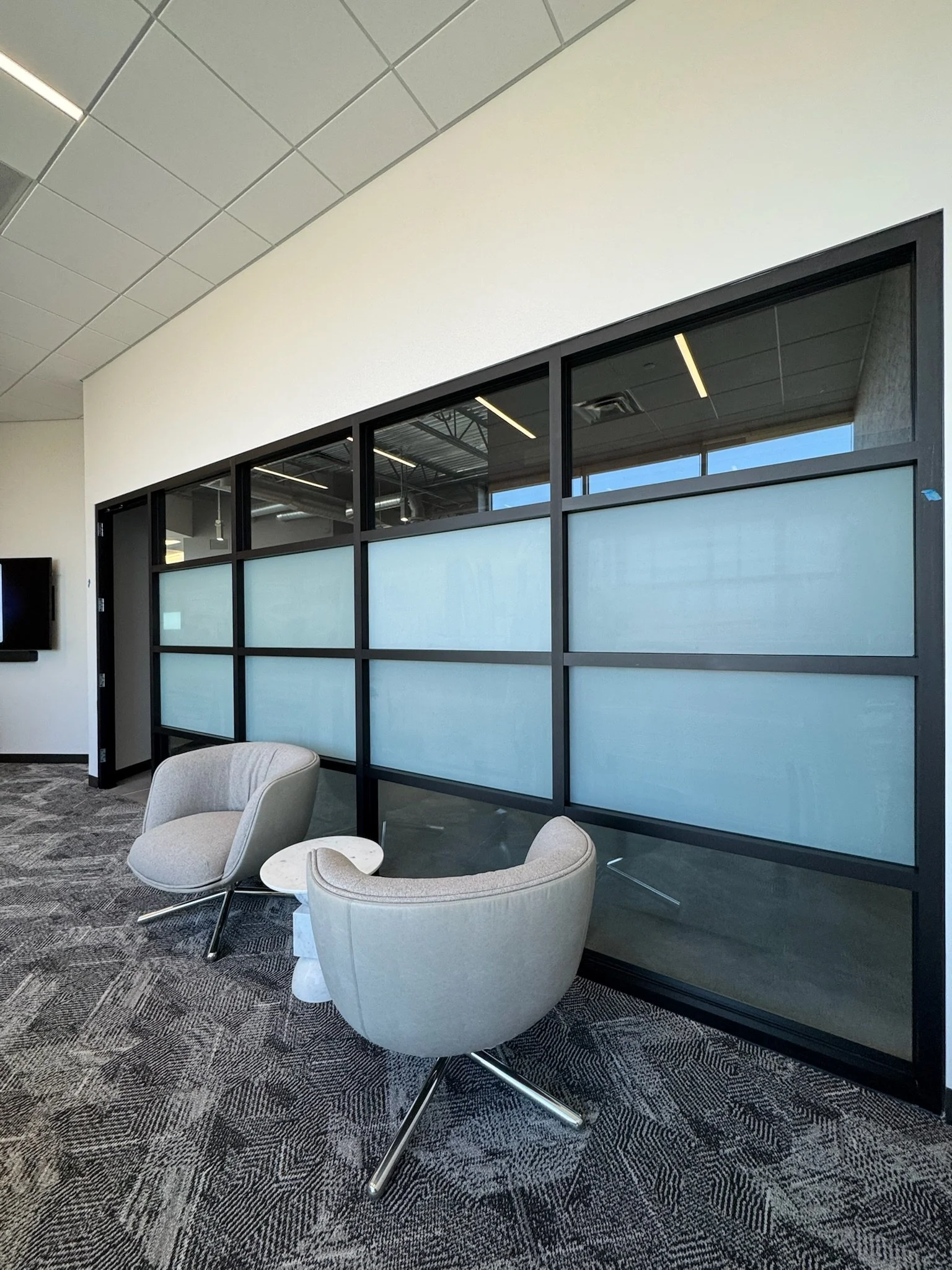 Modern office lounge area with beige armchairs, a small white table, a glass wall with black framing, and carpeted floor.