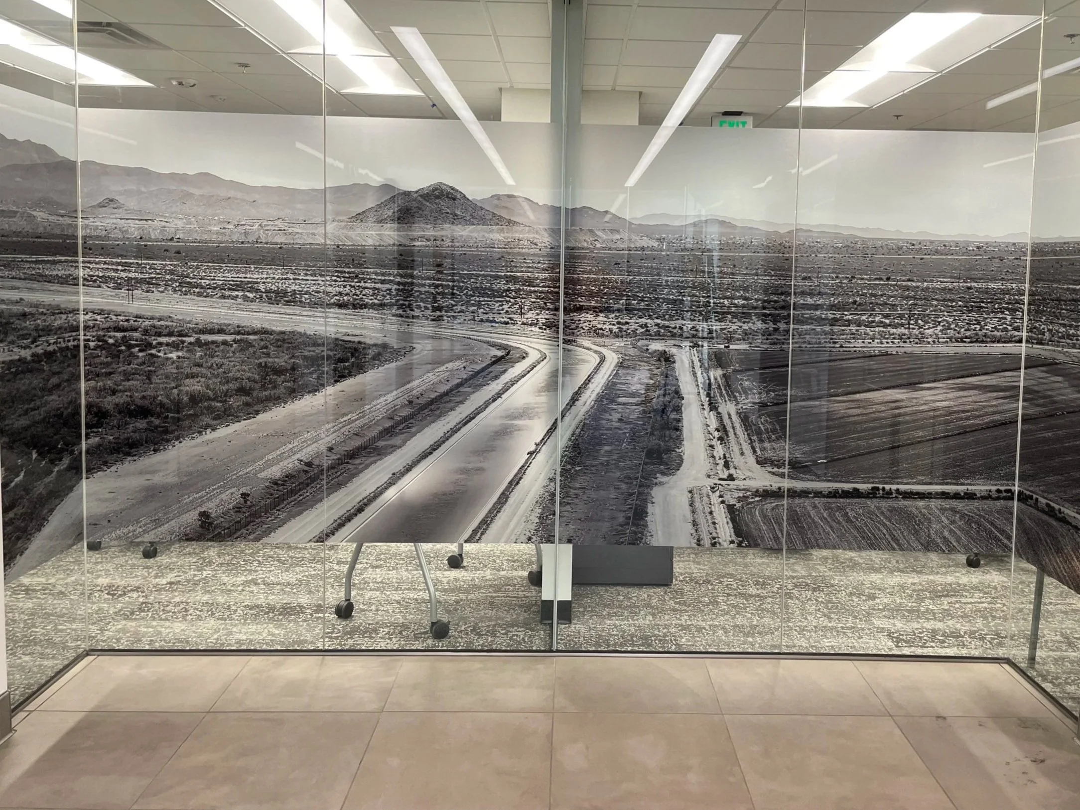 A large mural of a desert landscape with mountains in the background, displayed on glass panels inside a building. The mural appears to be printed on the panels, creating a continuous scene. The interior features a tiled ceiling and ceiling lights, with the reflection of room lights visible on the glass.