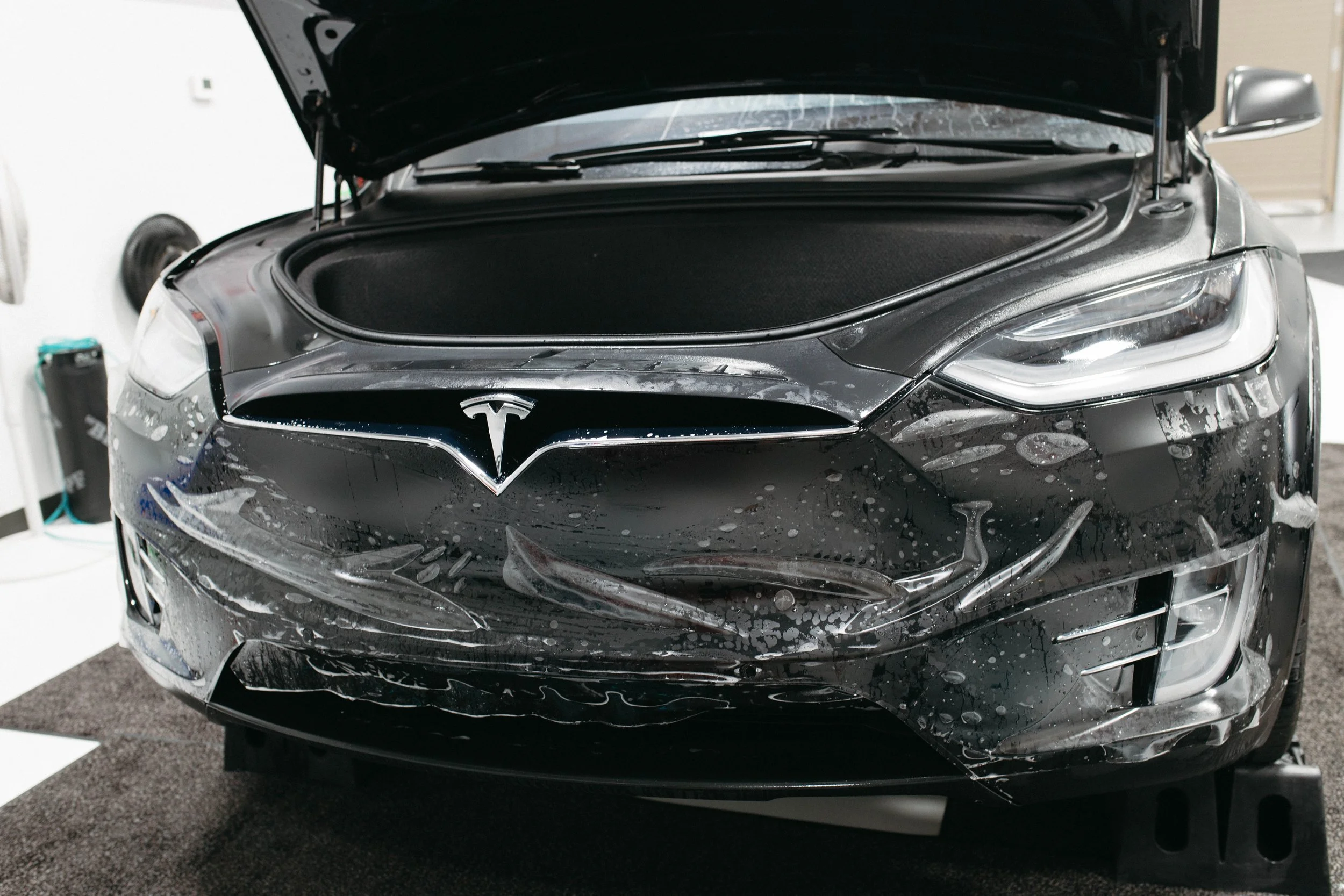 Close-up of a black Tesla car in a garage with bubbles and water on the front, hood open, and a garage door in the background.