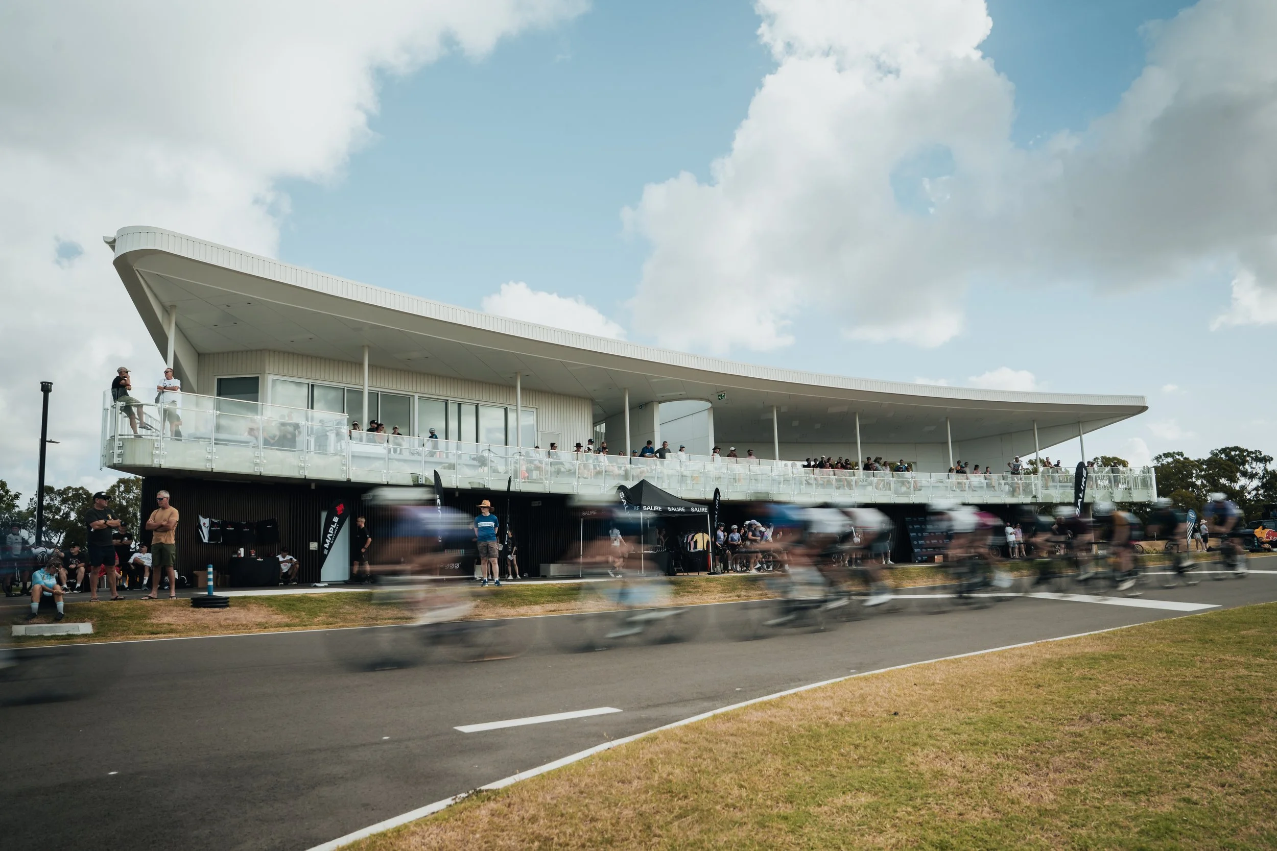 Cyclists racing on a track with a modern, white building and spectators watching from a balcony.