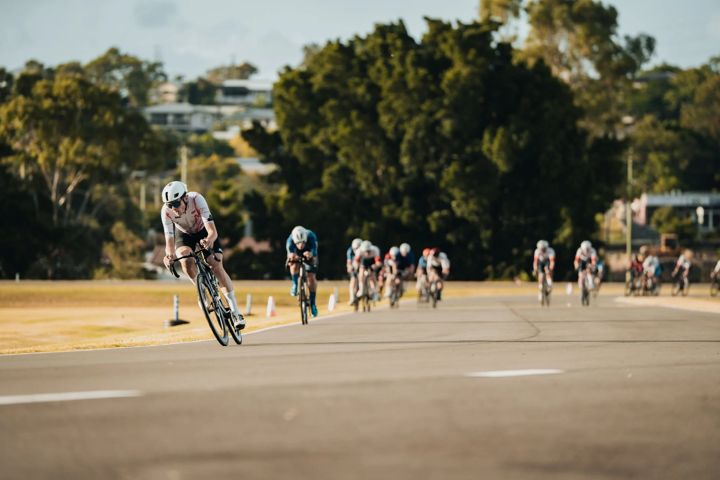 Cyclists participating in a bike race on a paved road with trees in the background and a group of cyclists in the distance.
