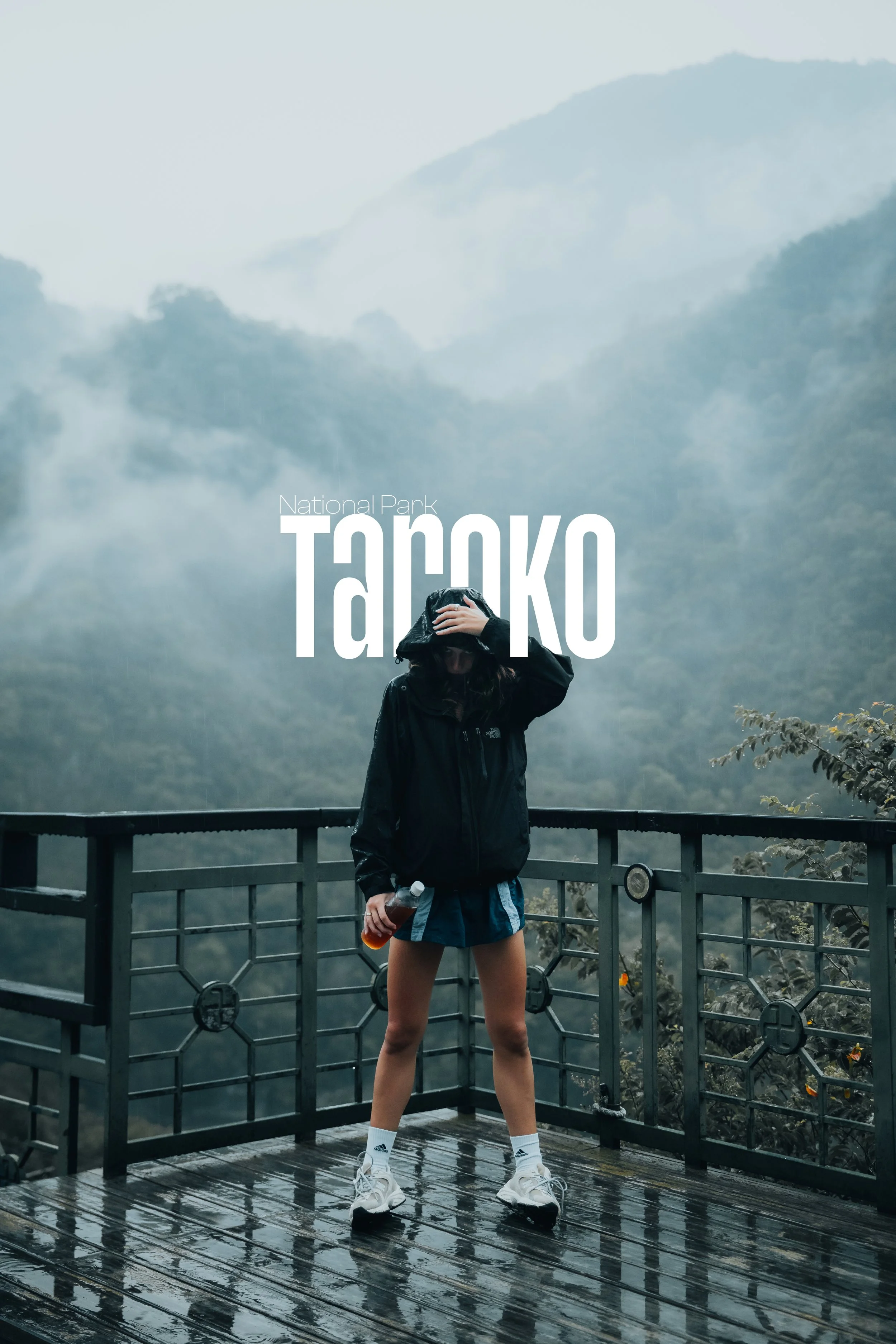 A person standing on a wet balcony in the rain, wearing a black jacket, shorts, and sneakers, with mountains and fog in the background in Taroko National Park.