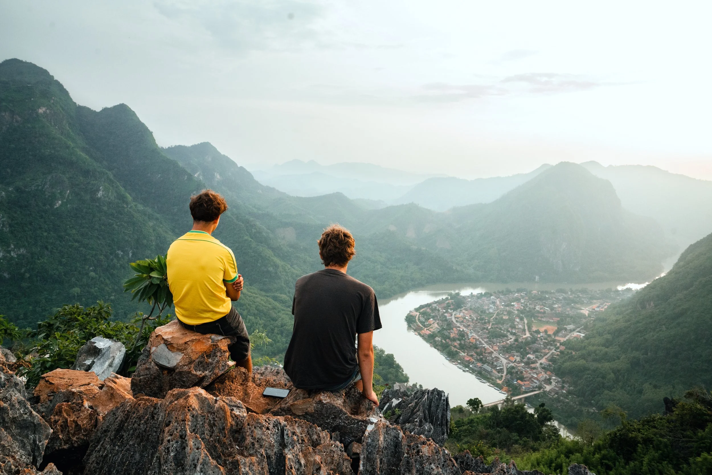Two young men sitting on rocks overlooking a lush green valley with a winding river and a small town below, surrounded by mountains.