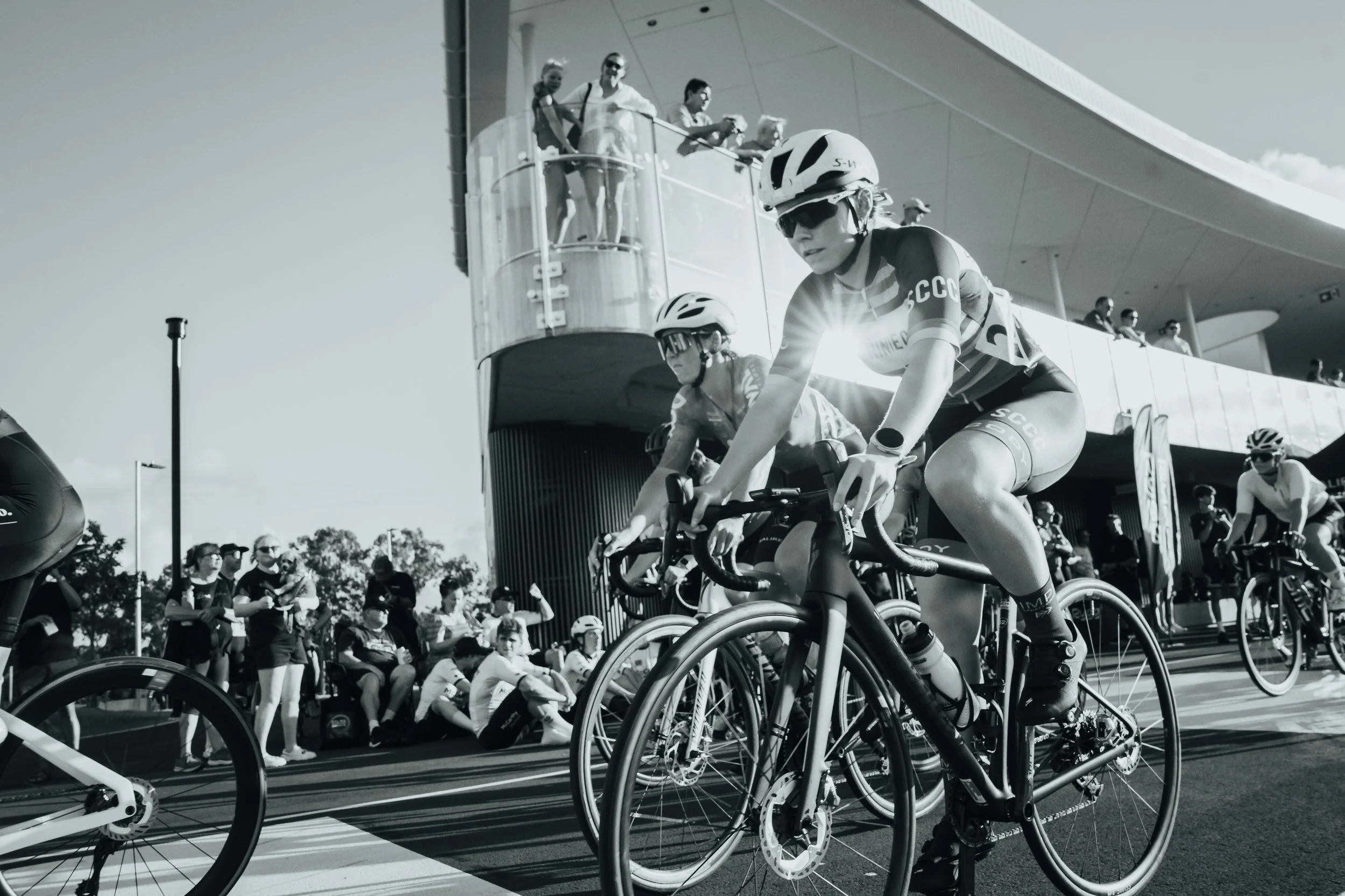 Cyclists in a race, wearing helmets and uniforms, riding on a track with spectators and a modern building in the background.