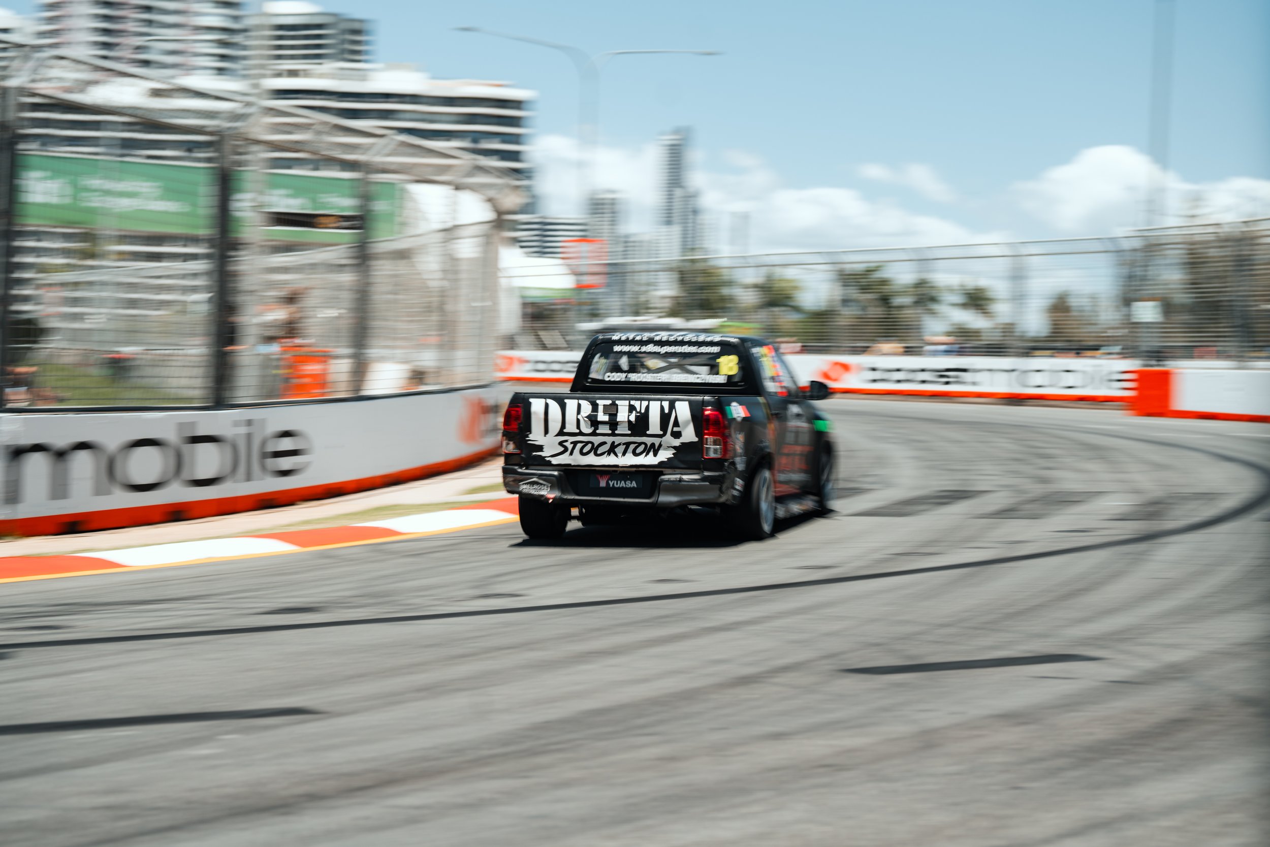 A black race truck with 'DRETA STOCKTON' written on the back, racing on a track near barriers with advertisements, in an urban area with tall buildings and blue skies overhead.