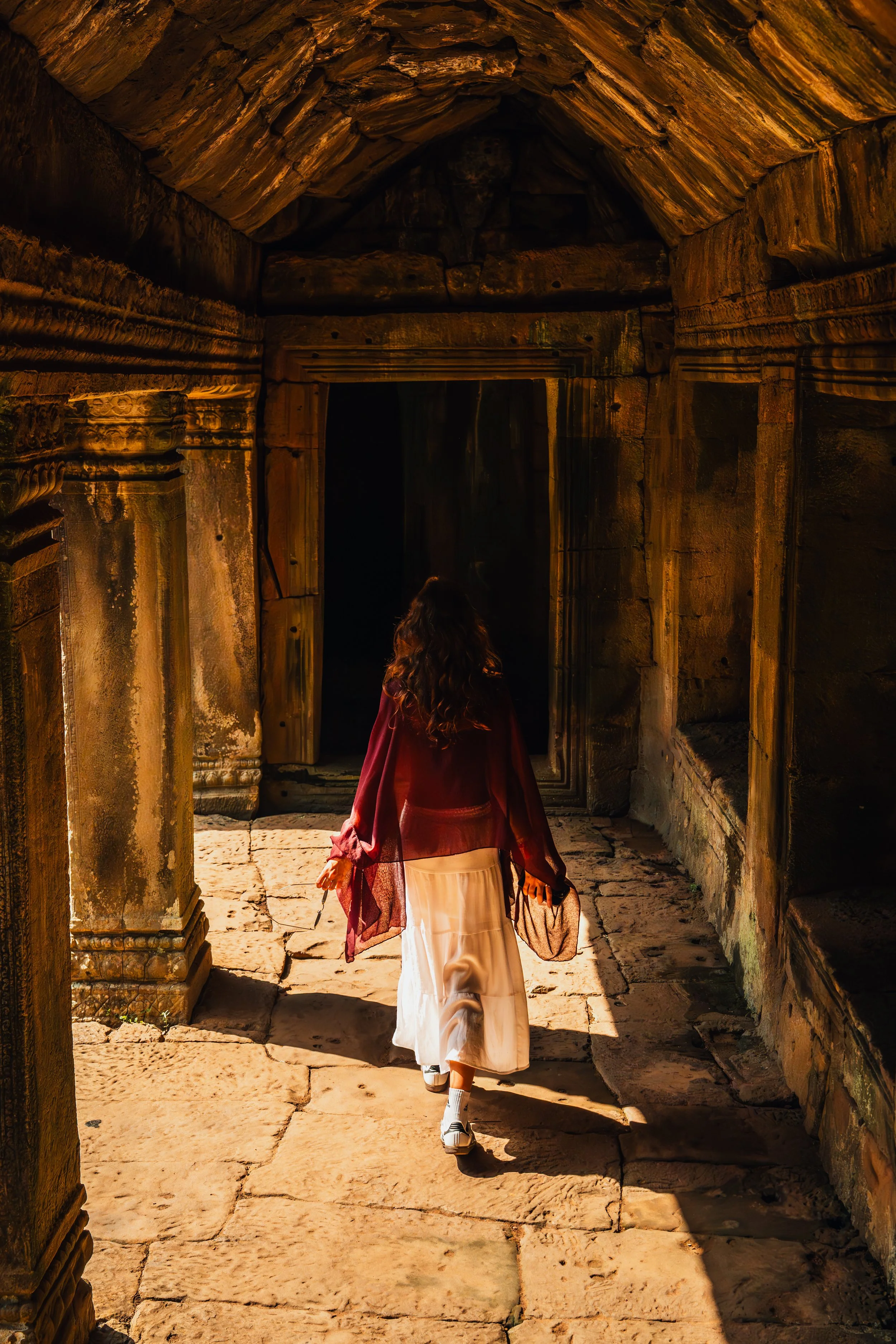 A person with long hair wearing a flowing white skirt, a burgundy top, and sneakers walking through ancient stone ruins at sunset, with warm sunlight casting long shadows.