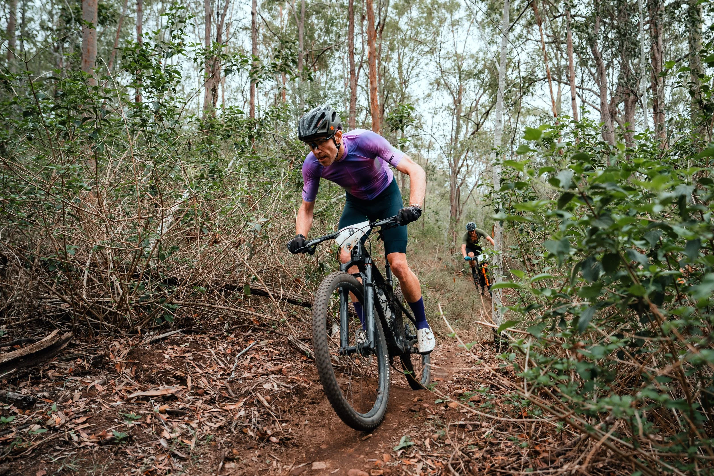 Two cyclists riding mountain bikes through a wooded trail, with one rider in the foreground and another in the background, surrounded by trees and shrubbery.