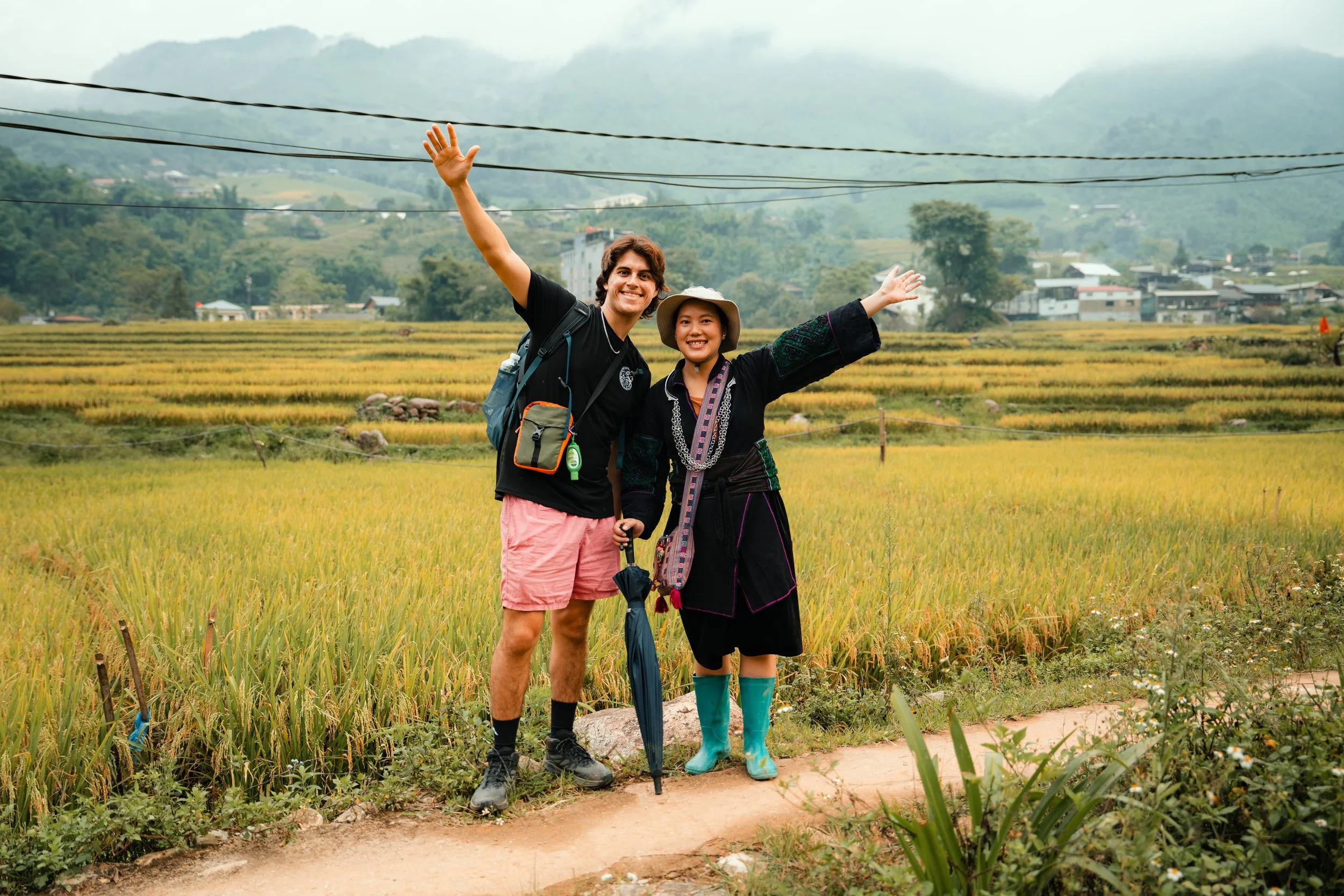 Two people standing on a dirt path in a lush green rice field, smiling and waving at the camera, with mountains and a cloudy sky in the background.