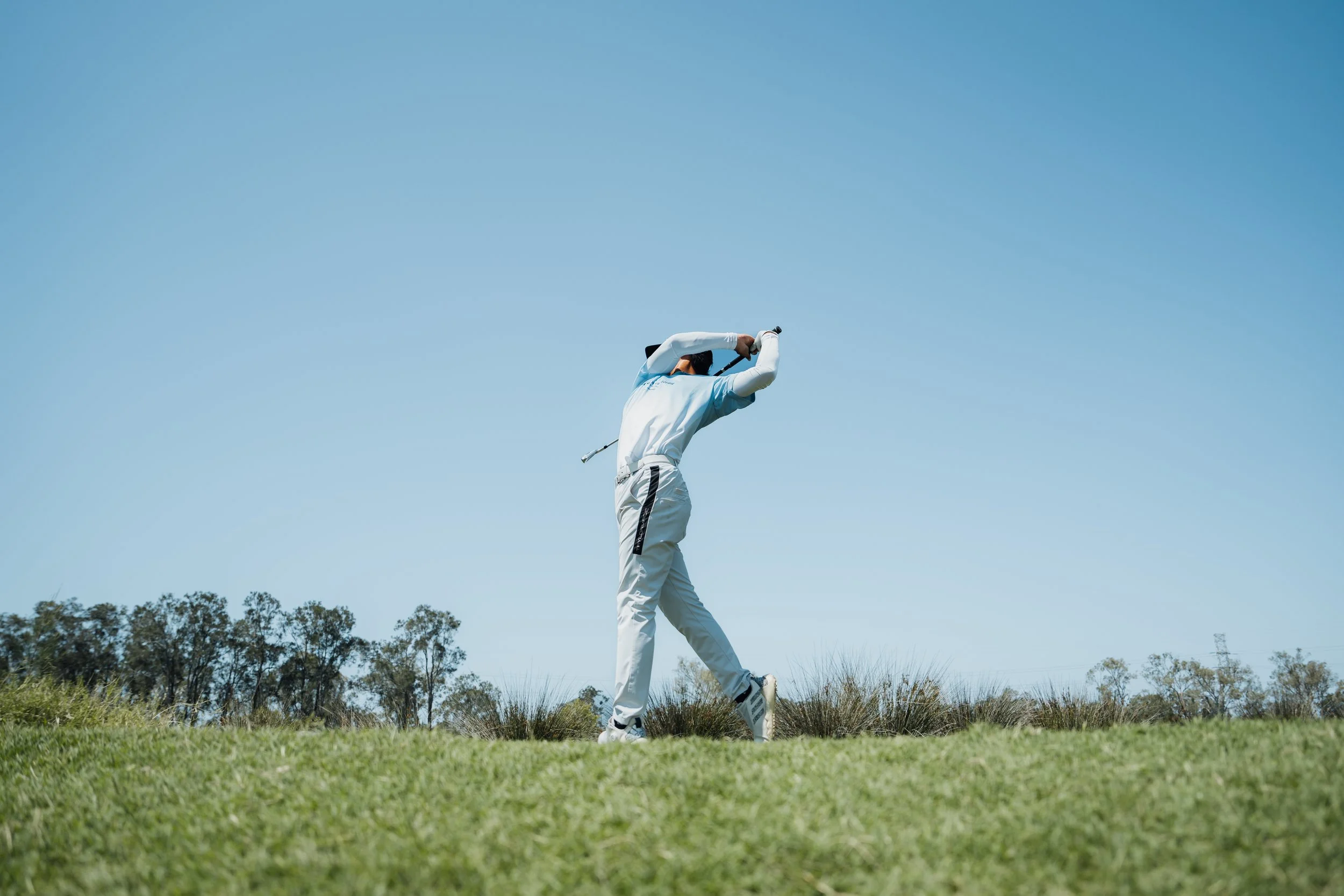 A golfer in white clothes swinging a golf club on a grassy golf course under a clear blue sky.
