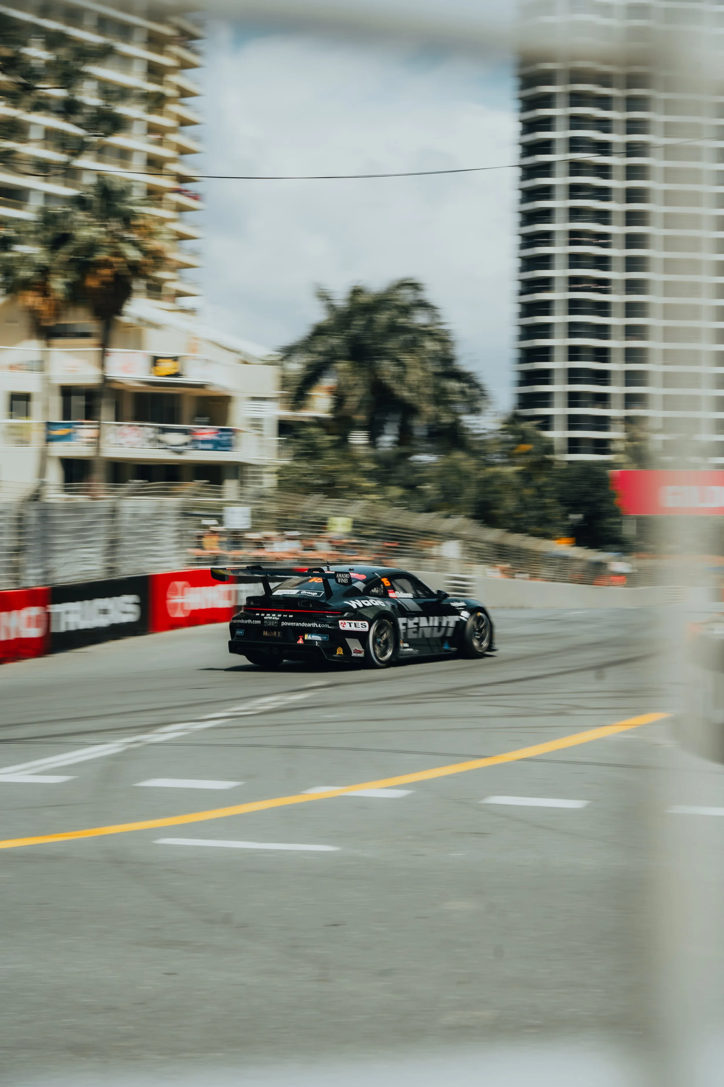 A black race car speeding on a city street during a race, with high-rise buildings and palm trees in the background.
