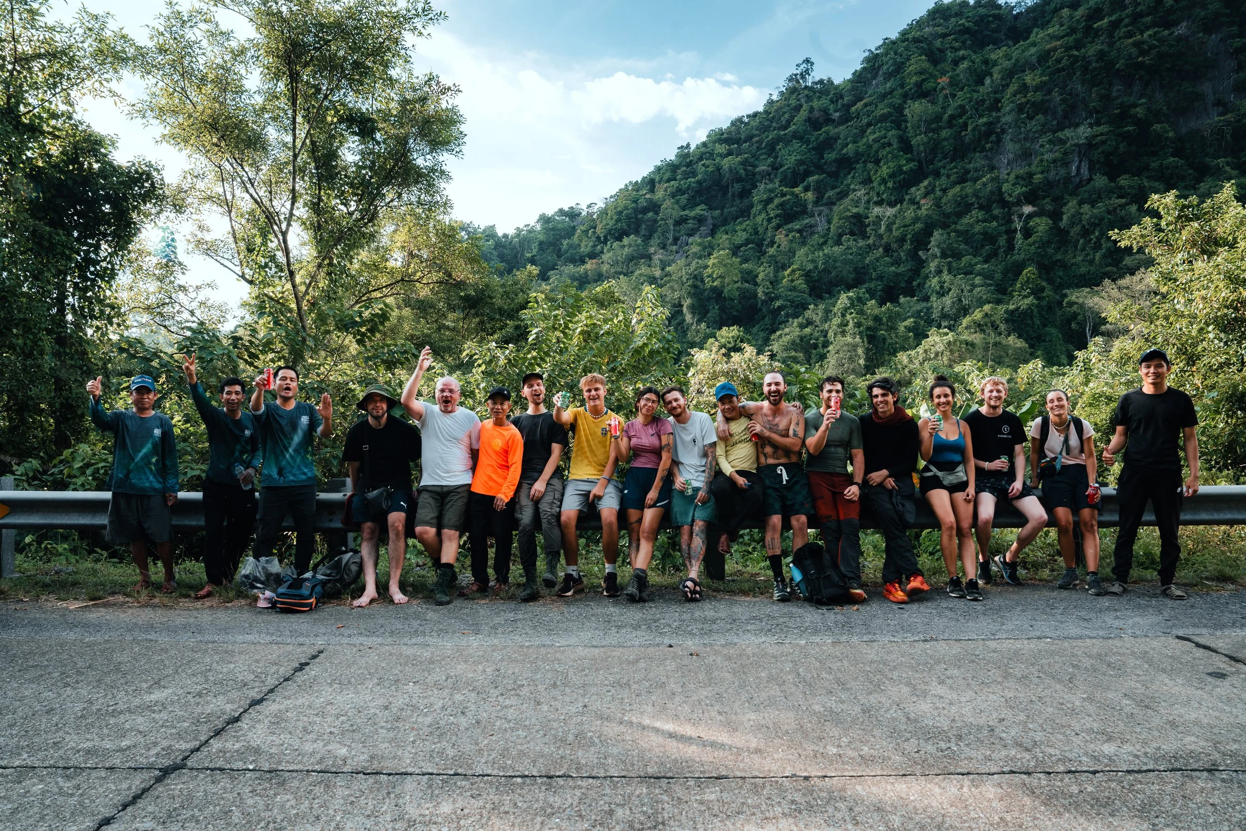 A group of people standing outdoors on the side of a road with lush green hills in the background. They are smiling, some making peace signs, and appear to be on a hike or outdoor adventure.
