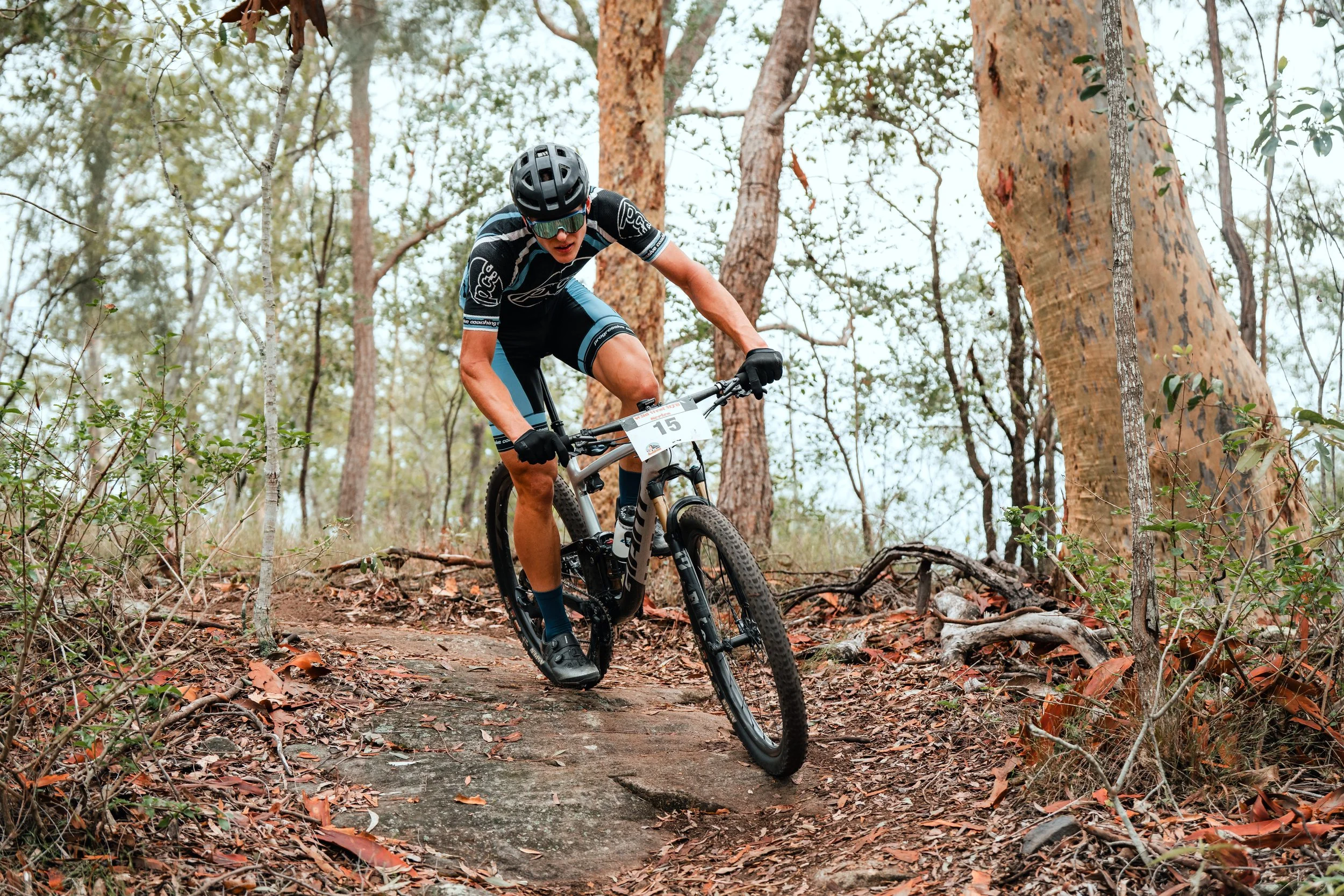 A man dressed in cycling gear riding a mountain bike on a dirt trail in a forest, wearing a helmet, sunglasses, and gloves.