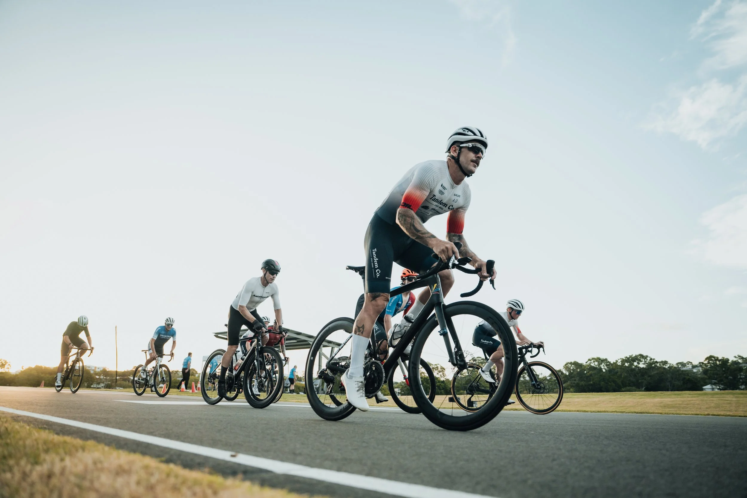 A group of cyclists riding on the road during daytime, with a clear sky and green trees in the background.