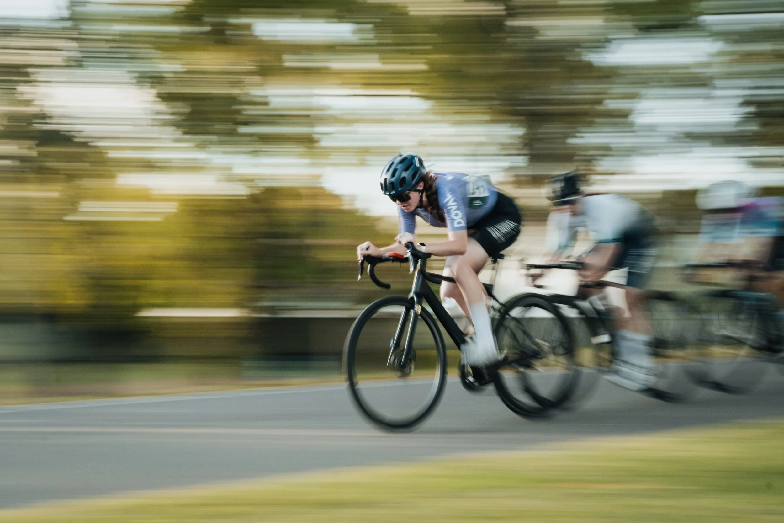 A cyclist wearing a blue helmet and blue jersey participating in a bike race on a paved road, with blurred background indicating high speed.