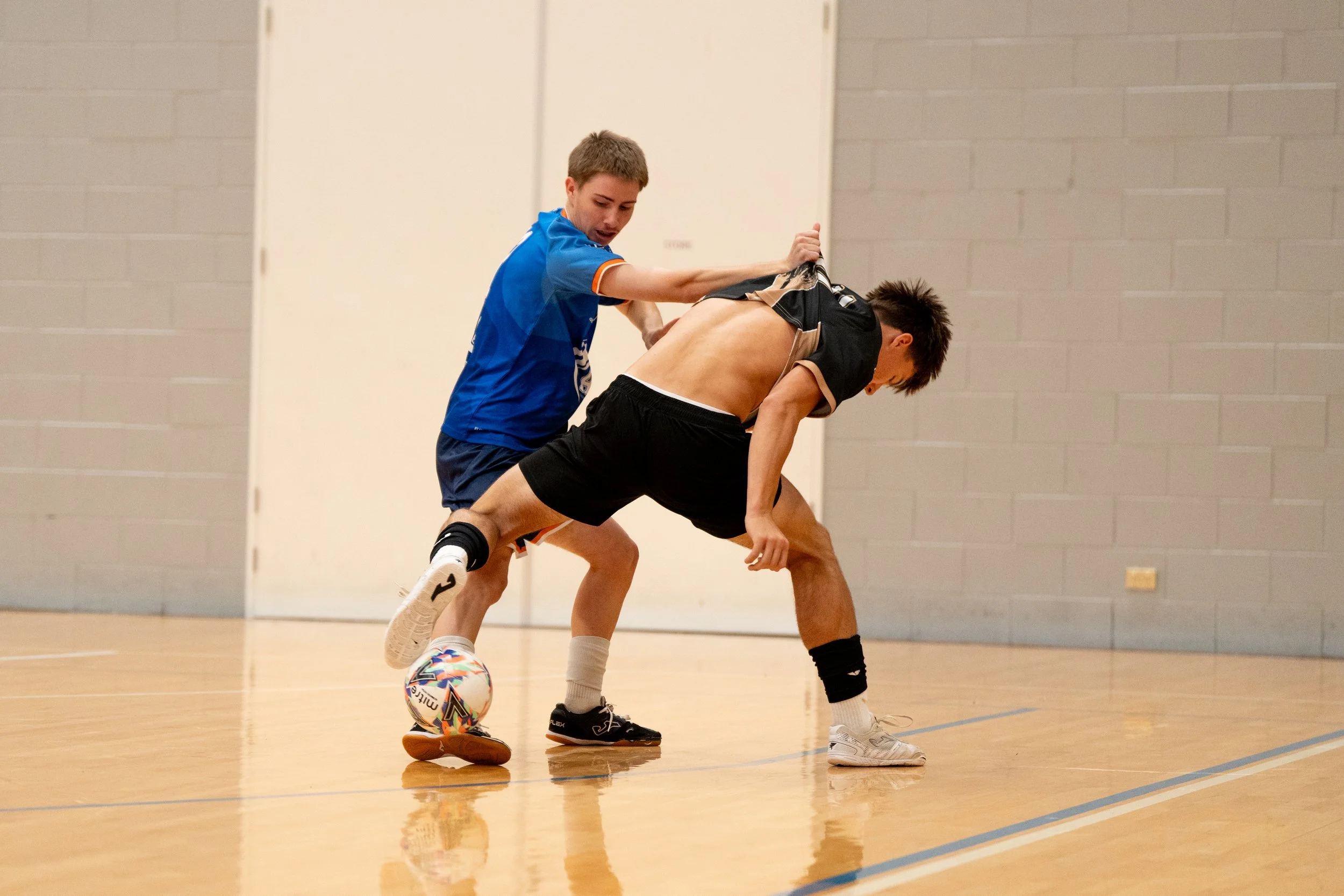 Two young men playing indoor soccer, one in a blue shirt and the other in black shorts, competing for the ball.