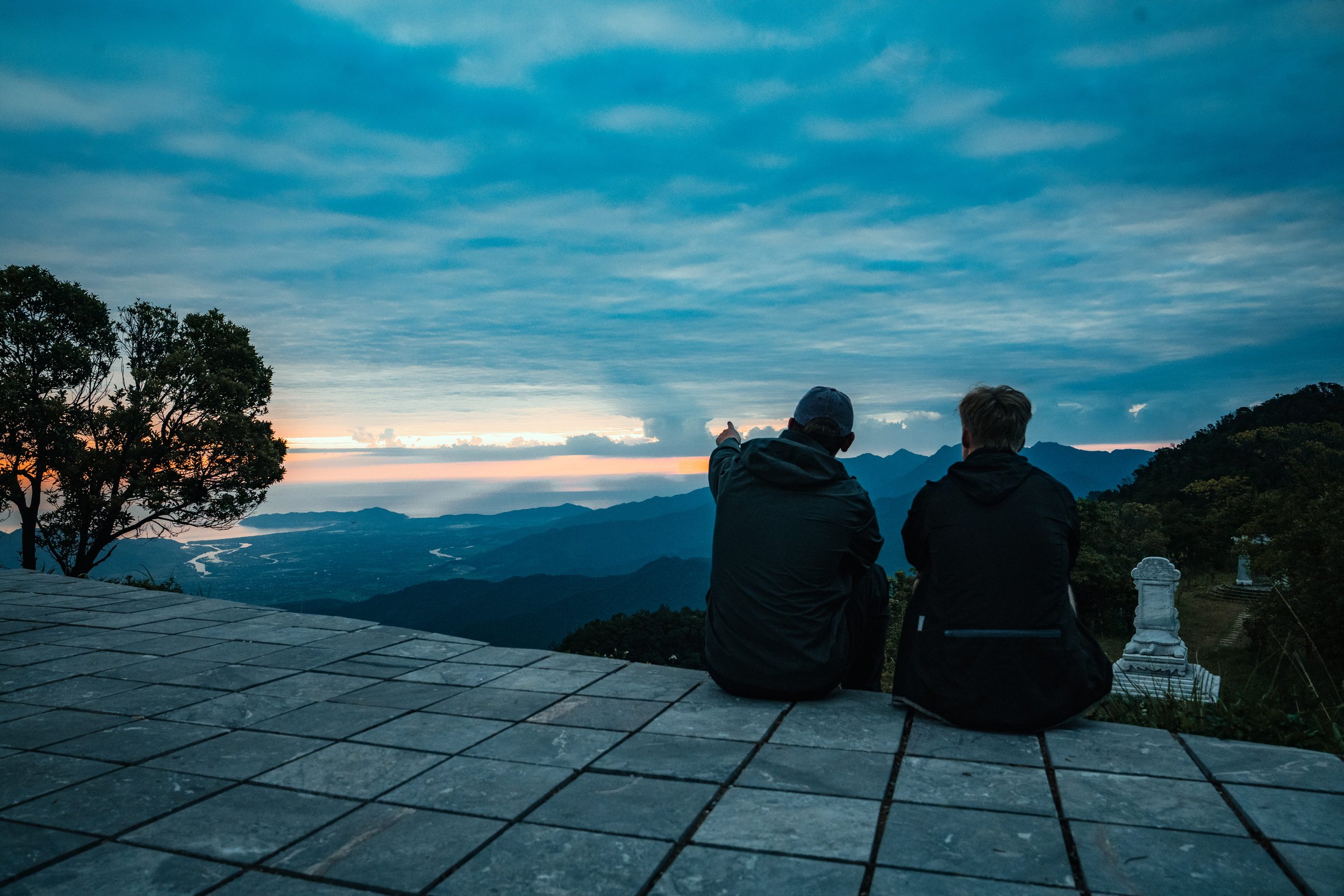 Two people sitting on a tiled platform overlooking a mountain landscape at sunset, one pointing towards the horizon.