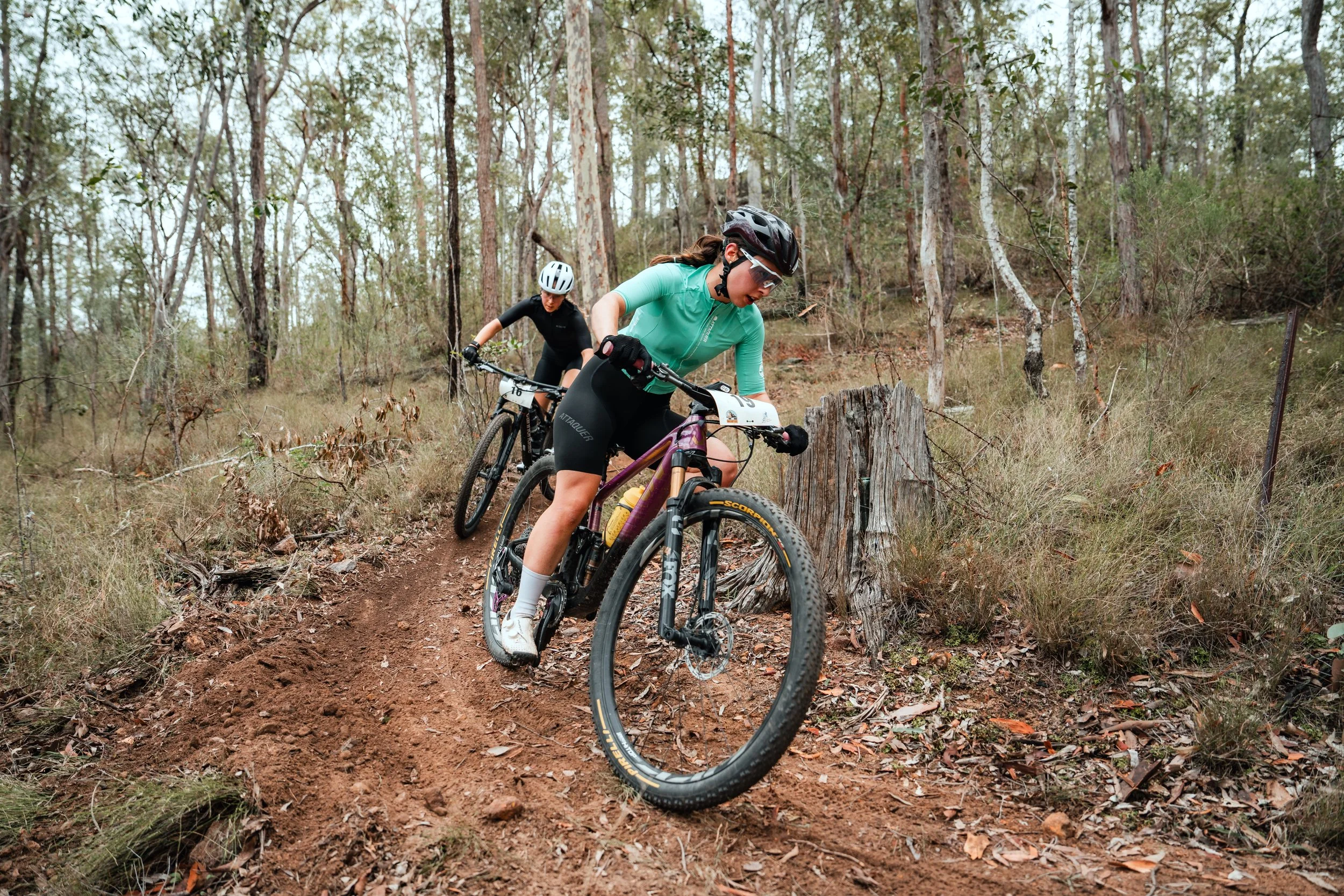 Two women mountain biking on a dirt trail through a wooded forest, wearing helmets and athletic clothing.