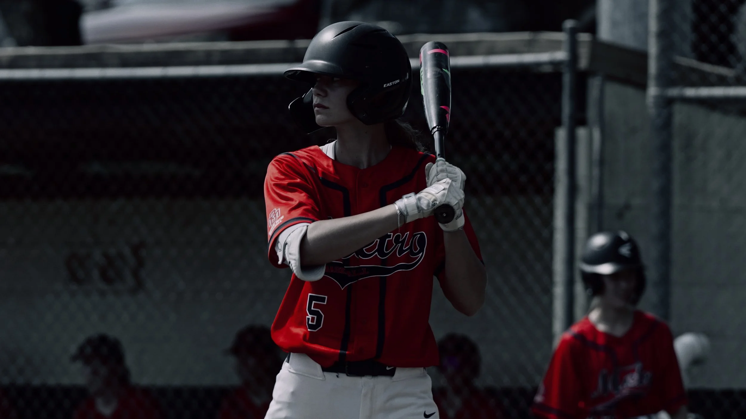 A female baseball player preparing to bat, wearing a black helmet, red jersey, and white pants, on a baseball field.