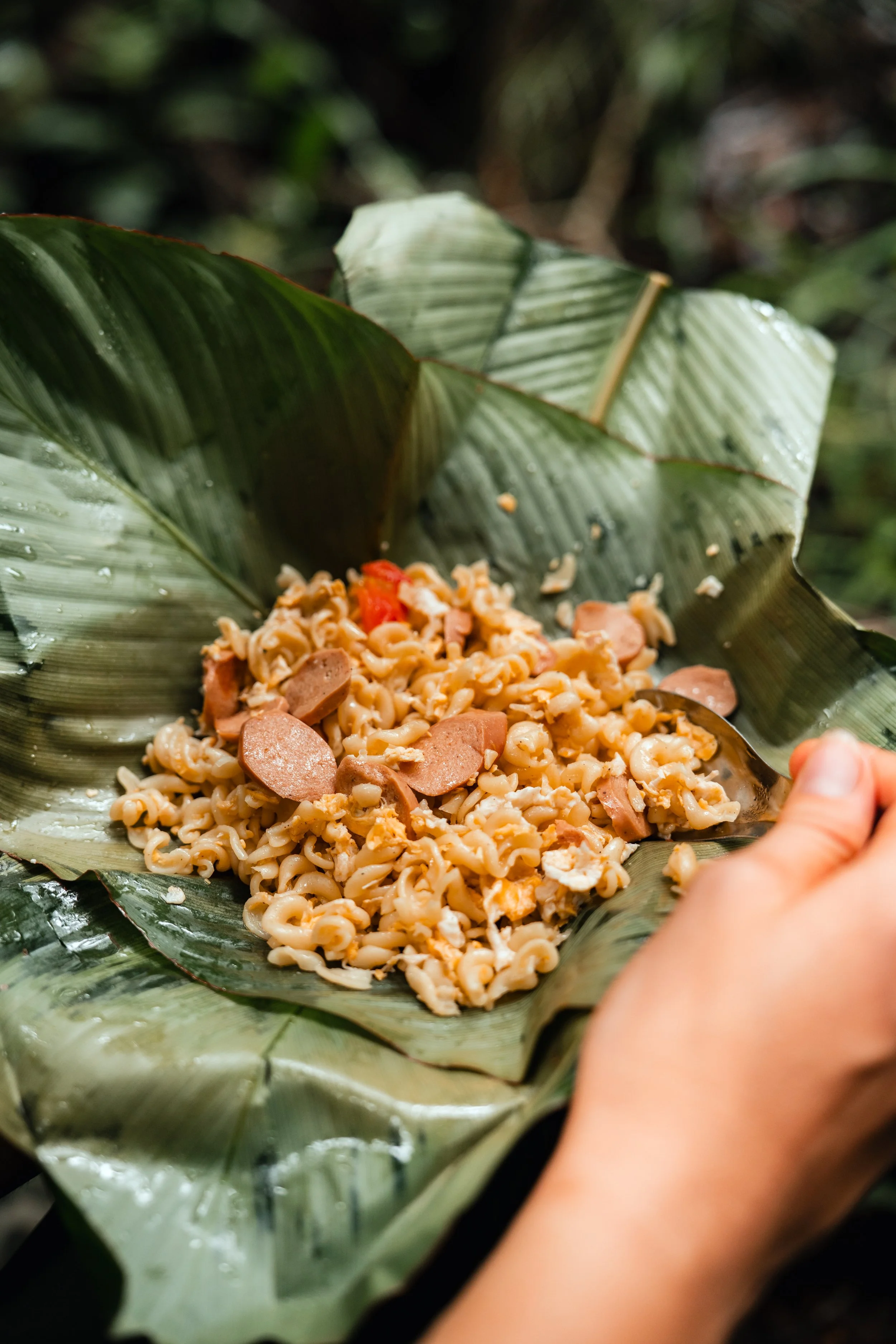 A person's hand holding a spoon, serving a dish of cooked instant ramen noodles with slices of sausage and bits of vegetables, all placed on banana leaves in a natural outdoor setting.