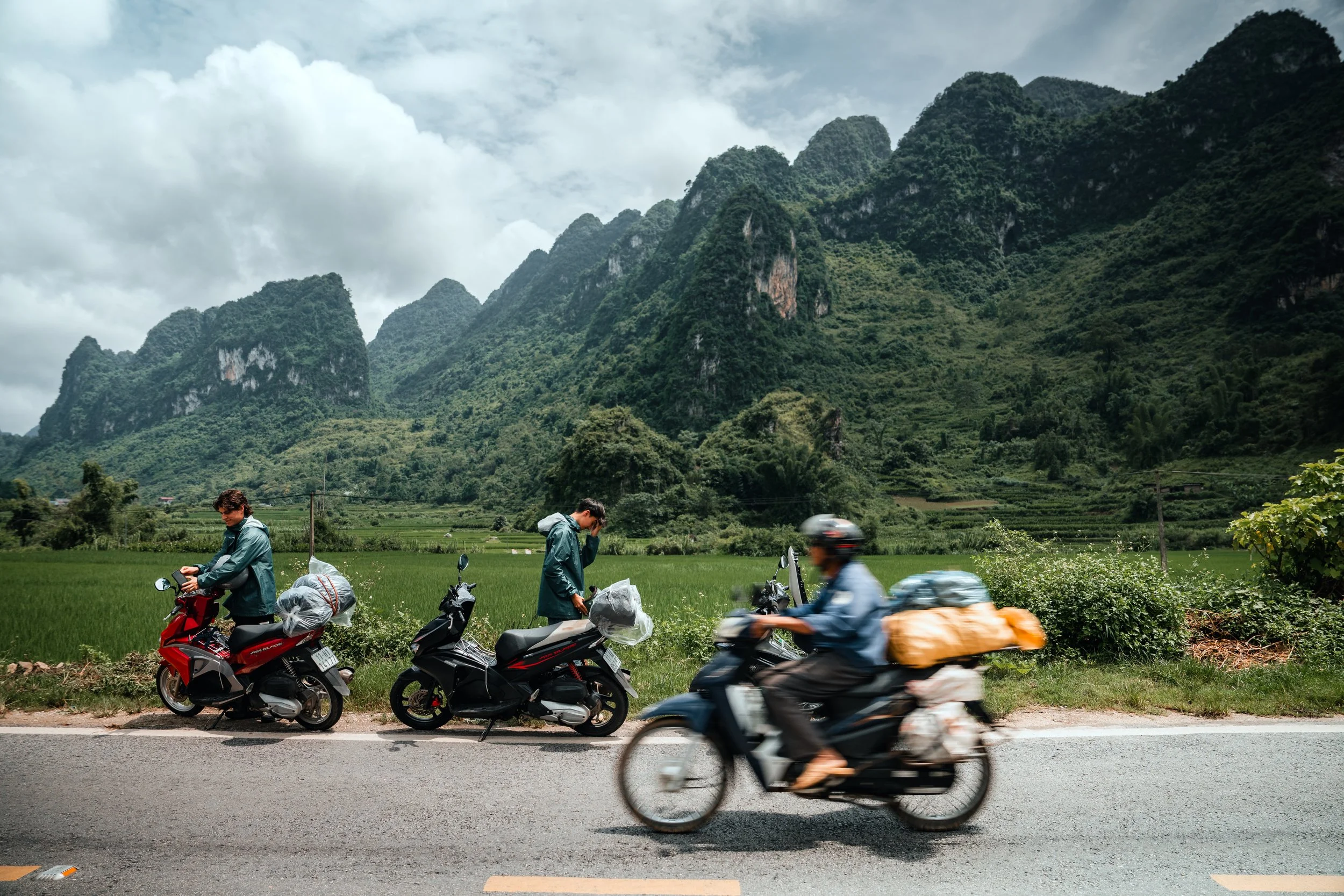 Two people standing next to parked scooters by a mountain landscape, with one person talking on the phone and the other looking at their phone, while a third person rides a motorcycle passing by on a paved road.