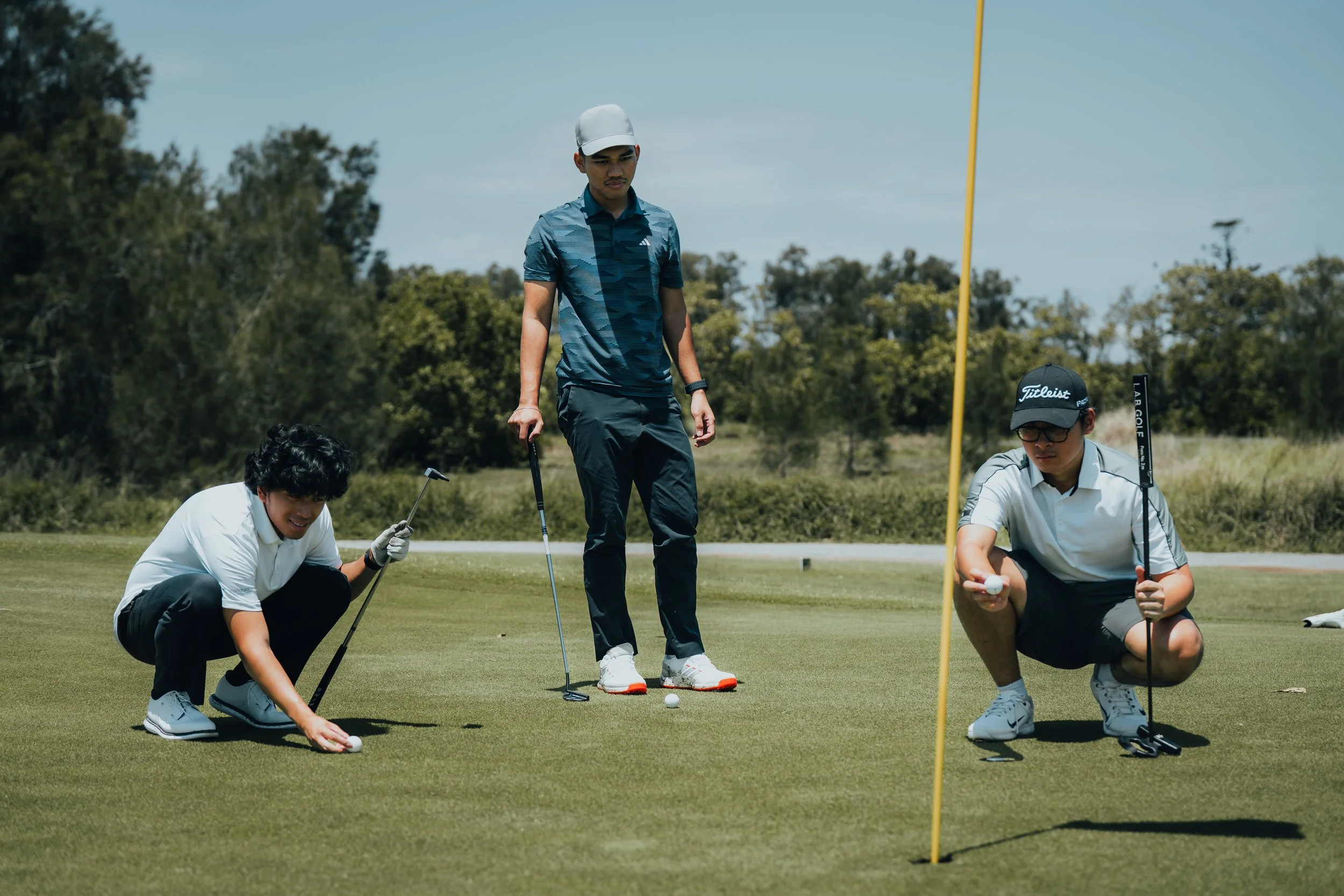 Three young men on a golf course, one standing and two squatting handling golf balls, with a flagstick nearby, trees and a blue sky in the background.