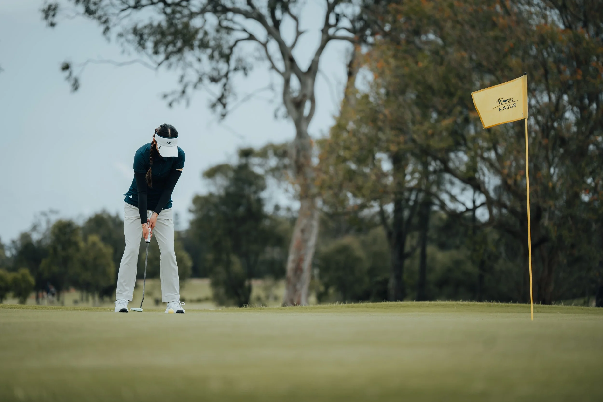 A female golfer prepares to putt on a golf course during daytime, with a yellow flag marked 'BULKA' waving in the background and trees in the distance.