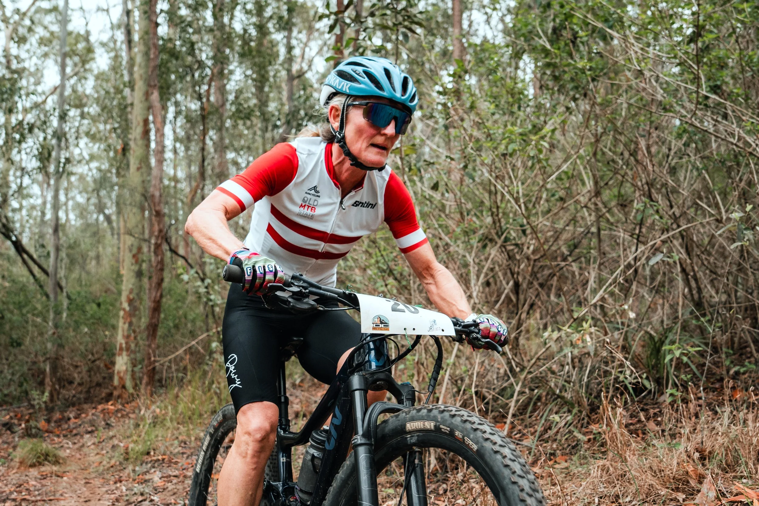 An older woman mountain biking through a forest trail, wearing a blue helmet, sunglasses, a white and red cycling jersey, black shorts, and colorful gloves.