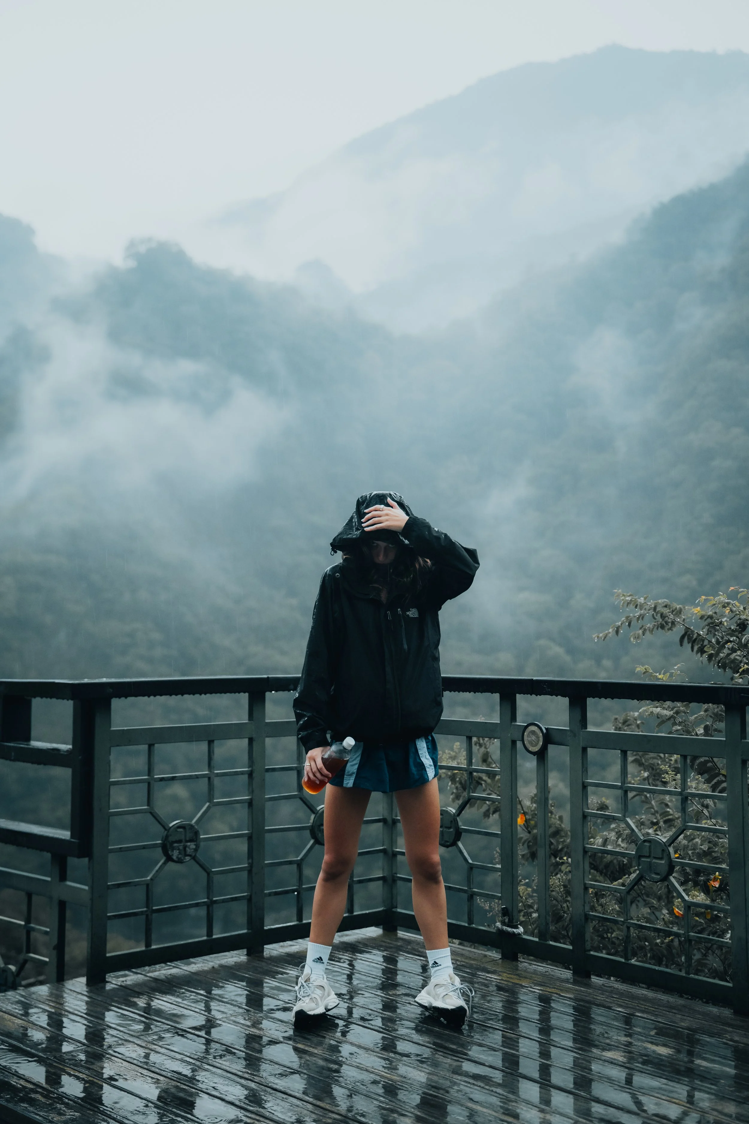 A person wearing a black rain jacket, shorts, and sneakers standing on a platform in the rain, holding a water bottle, with a foggy mountain landscape in the background.
