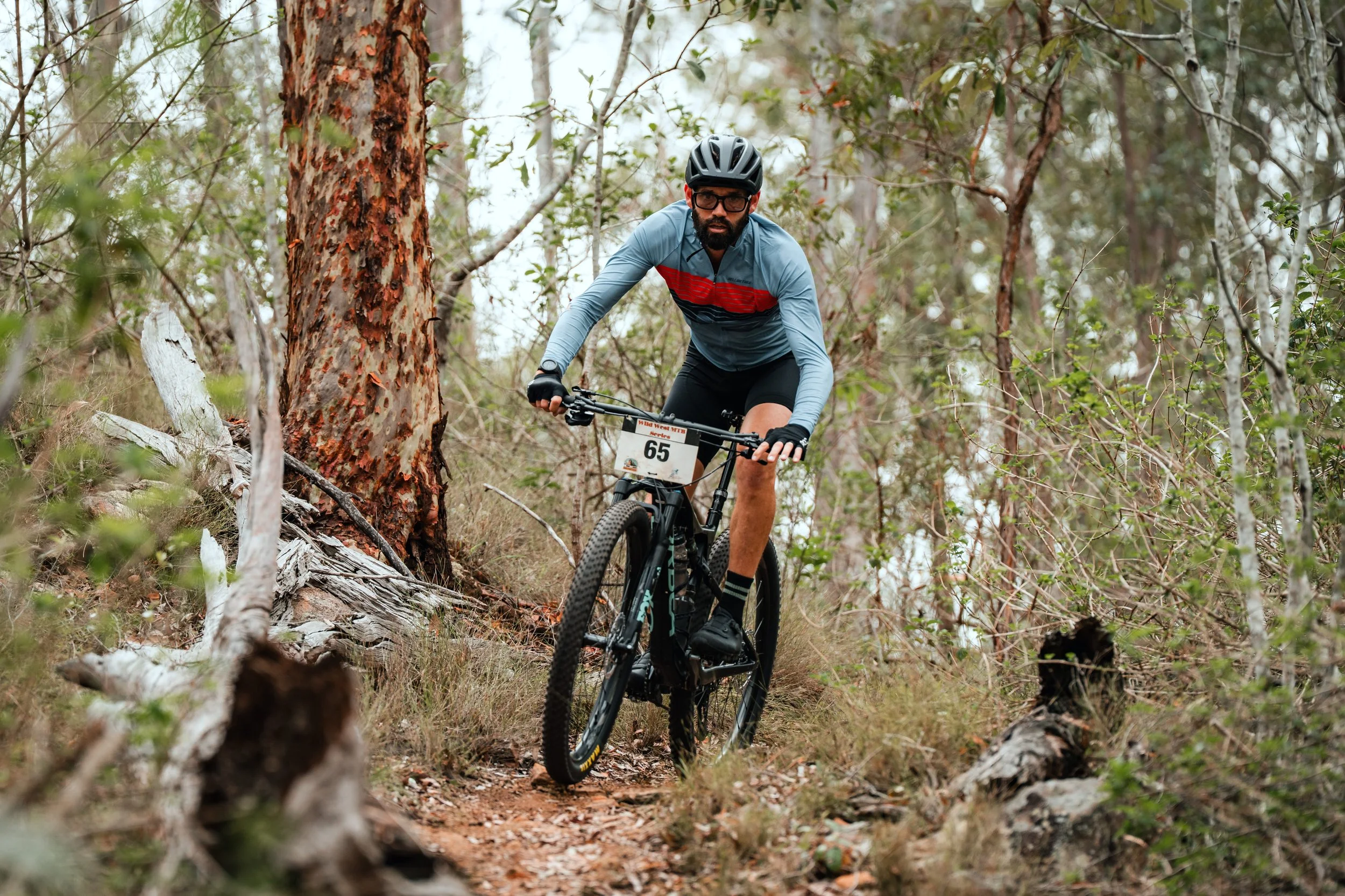A man wearing a helmet, glasses, a blue long-sleeve shirt with red stripes, black shorts, and gloves riding a mountain bike on a narrow dirt trail through a wooded area with trees and shrubs.