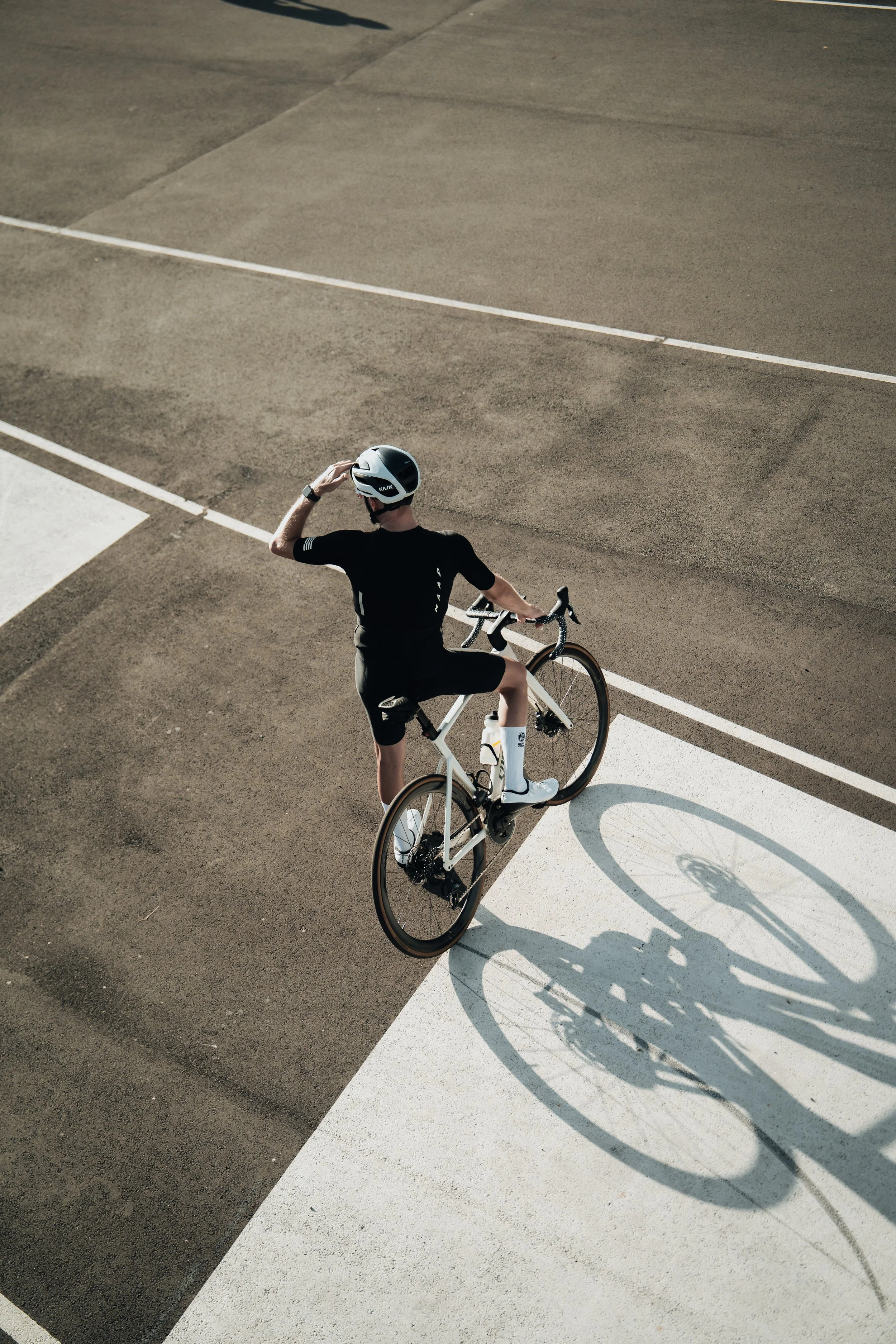A cyclist in black gear and a helmet standing with one foot on a white road bike on a paved surface, casting a shadow.