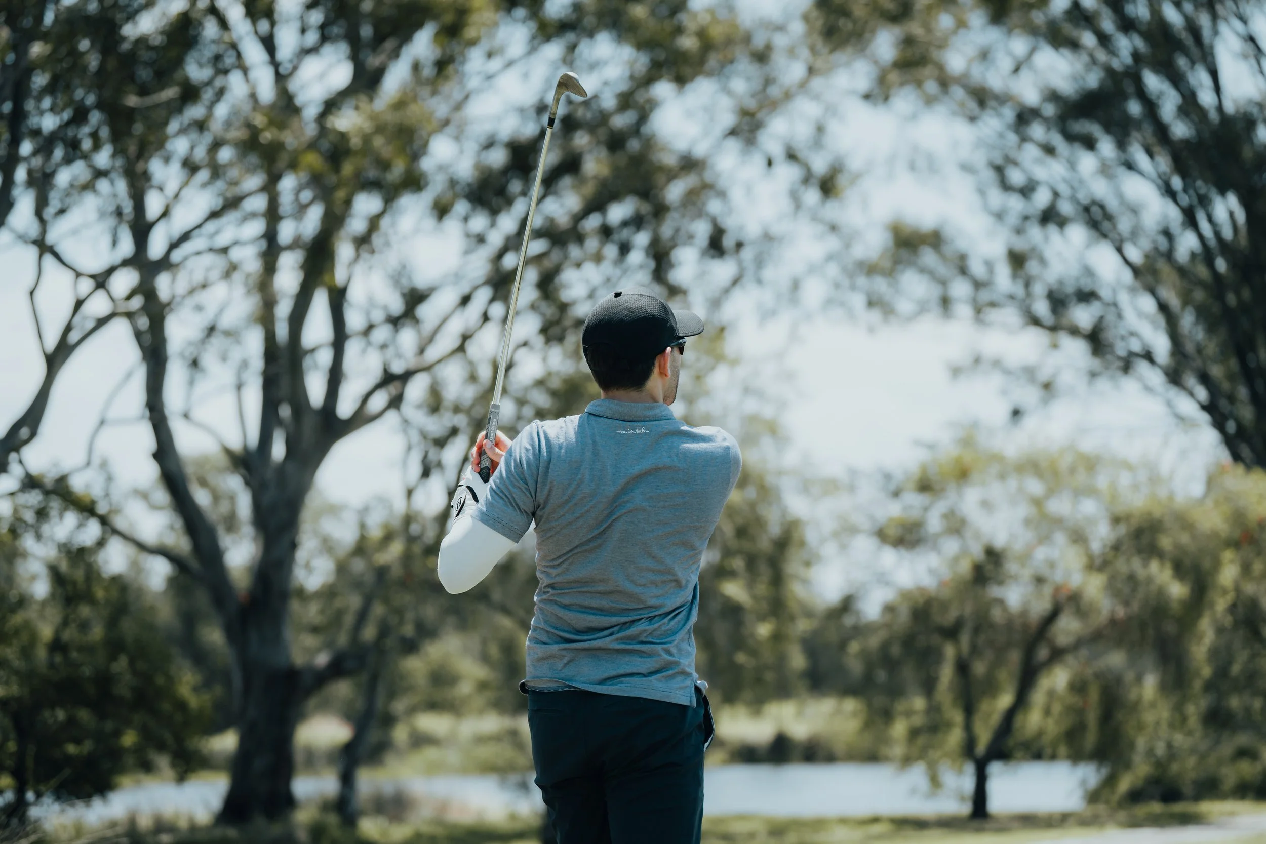 A man playing golf outdoors, swinging a golf club near a water hazard with trees in the background.