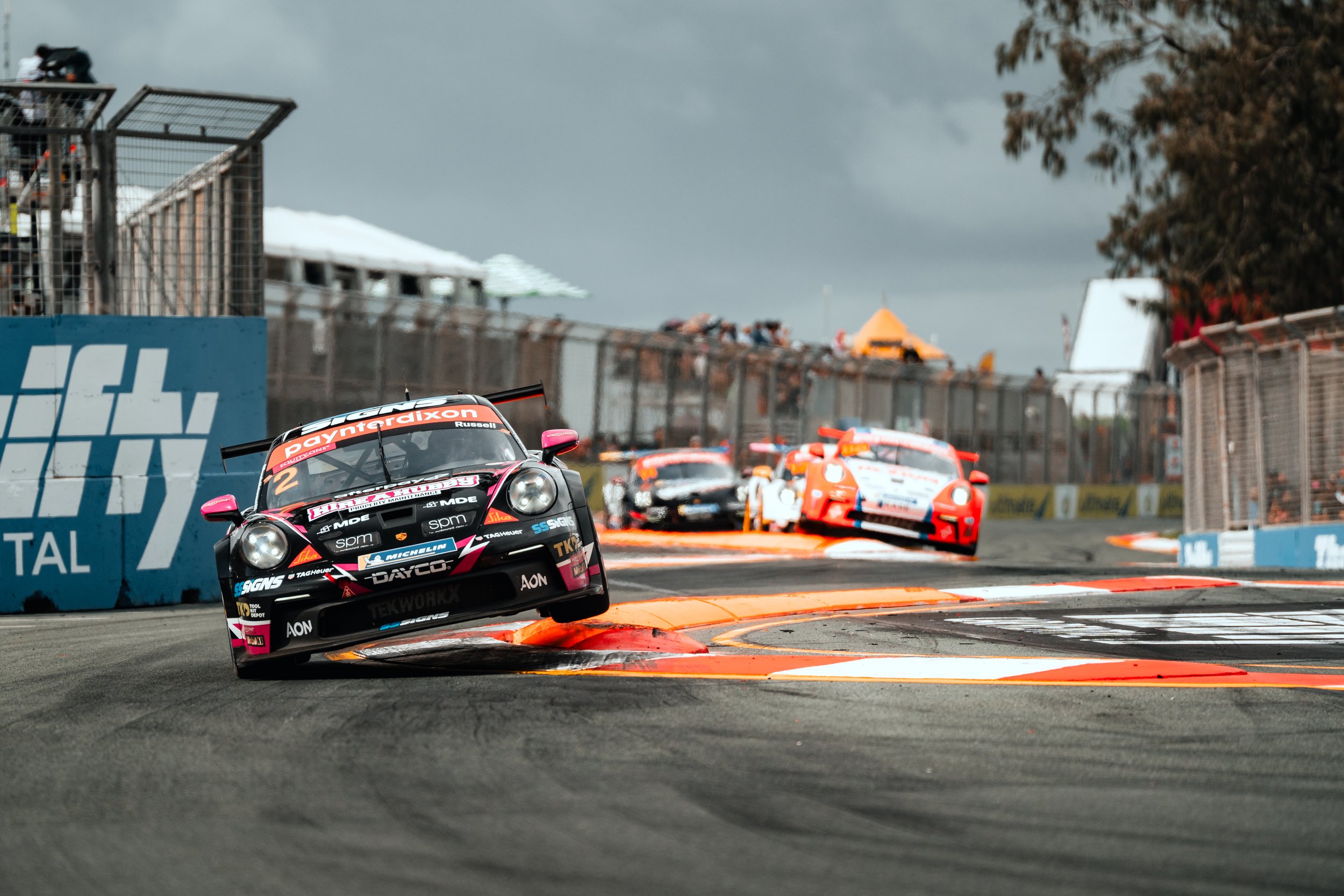 Race cars speeding around a track during a motorsport event, with the front car lifted off the ground as it navigates a corner, and multiple cars following behind on a cloudy day.