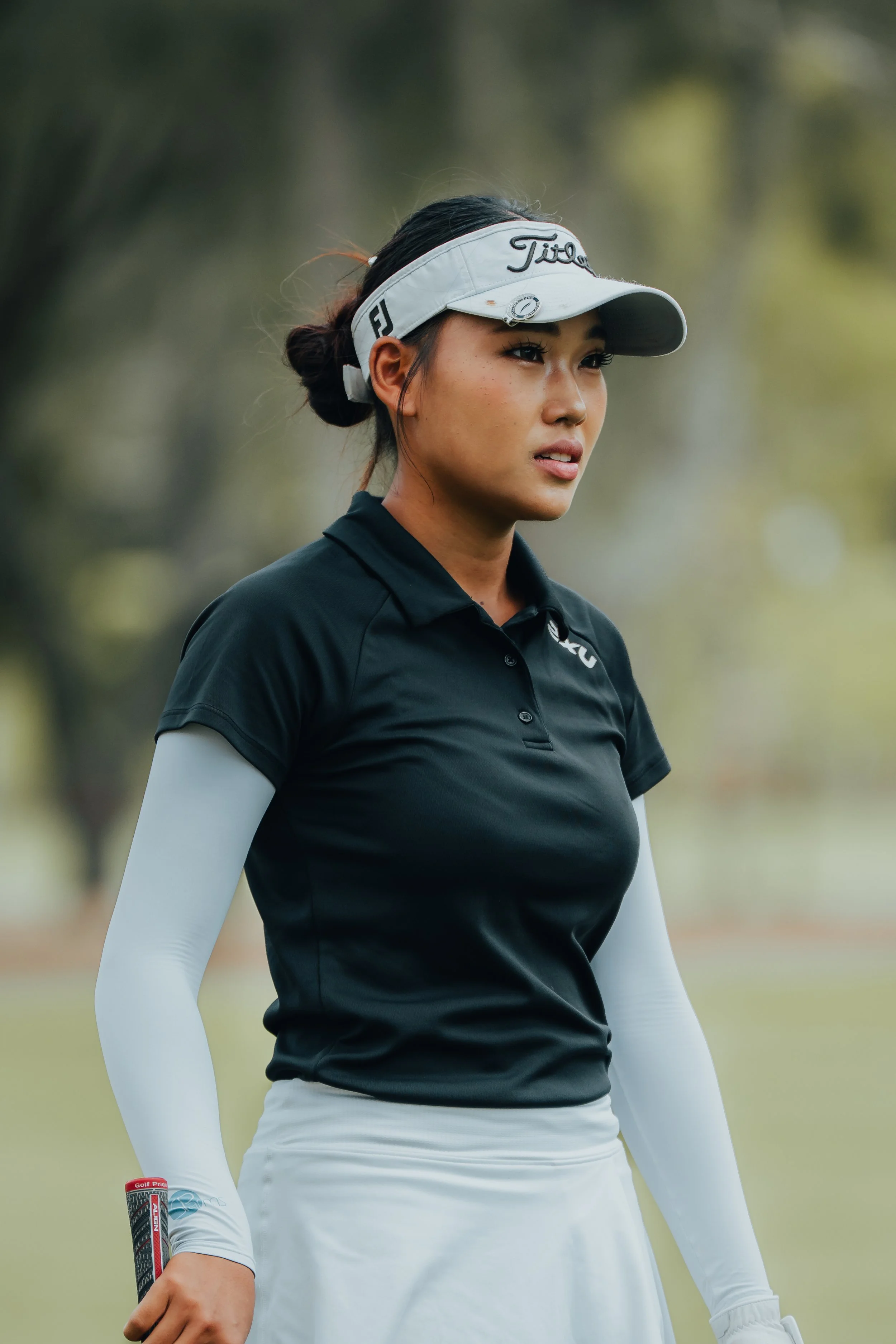 A female golfer wearing a white Titleist visor, black polo shirt, and white sleeve covers, holding a golf club on a golf course.