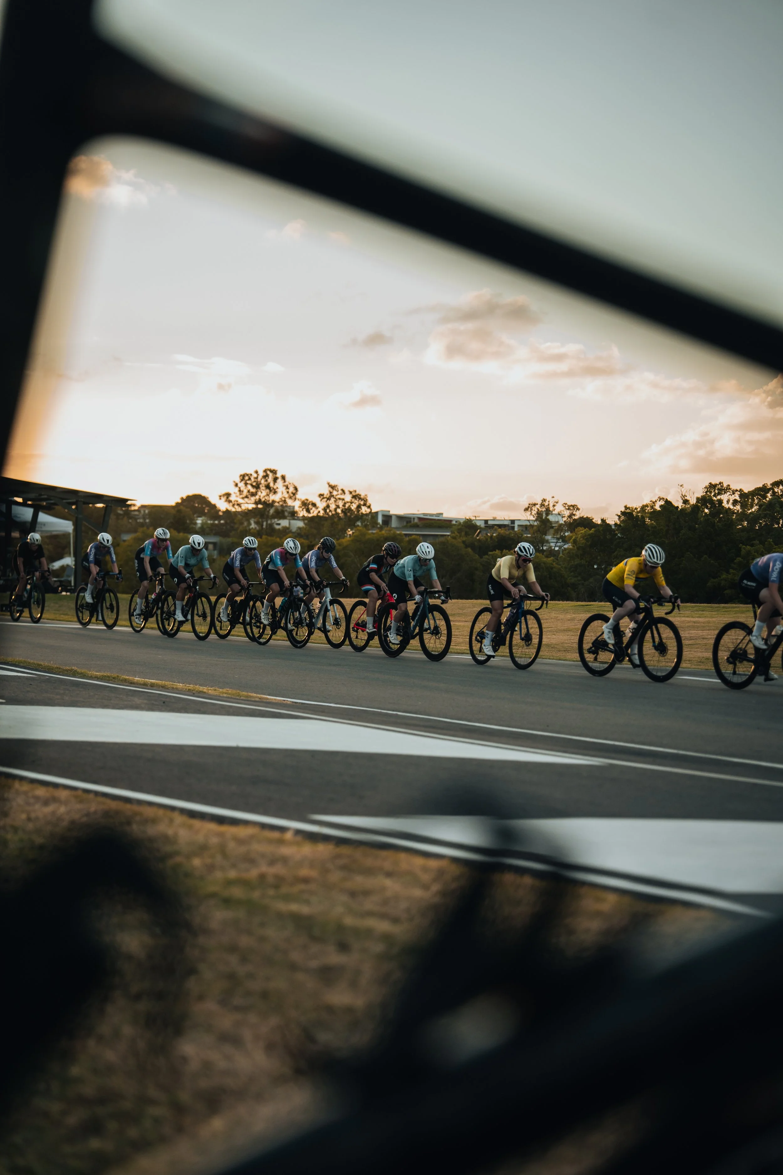 A group of cyclists riding on a track at sunset, seen through a slightly blurred view from behind a fence.